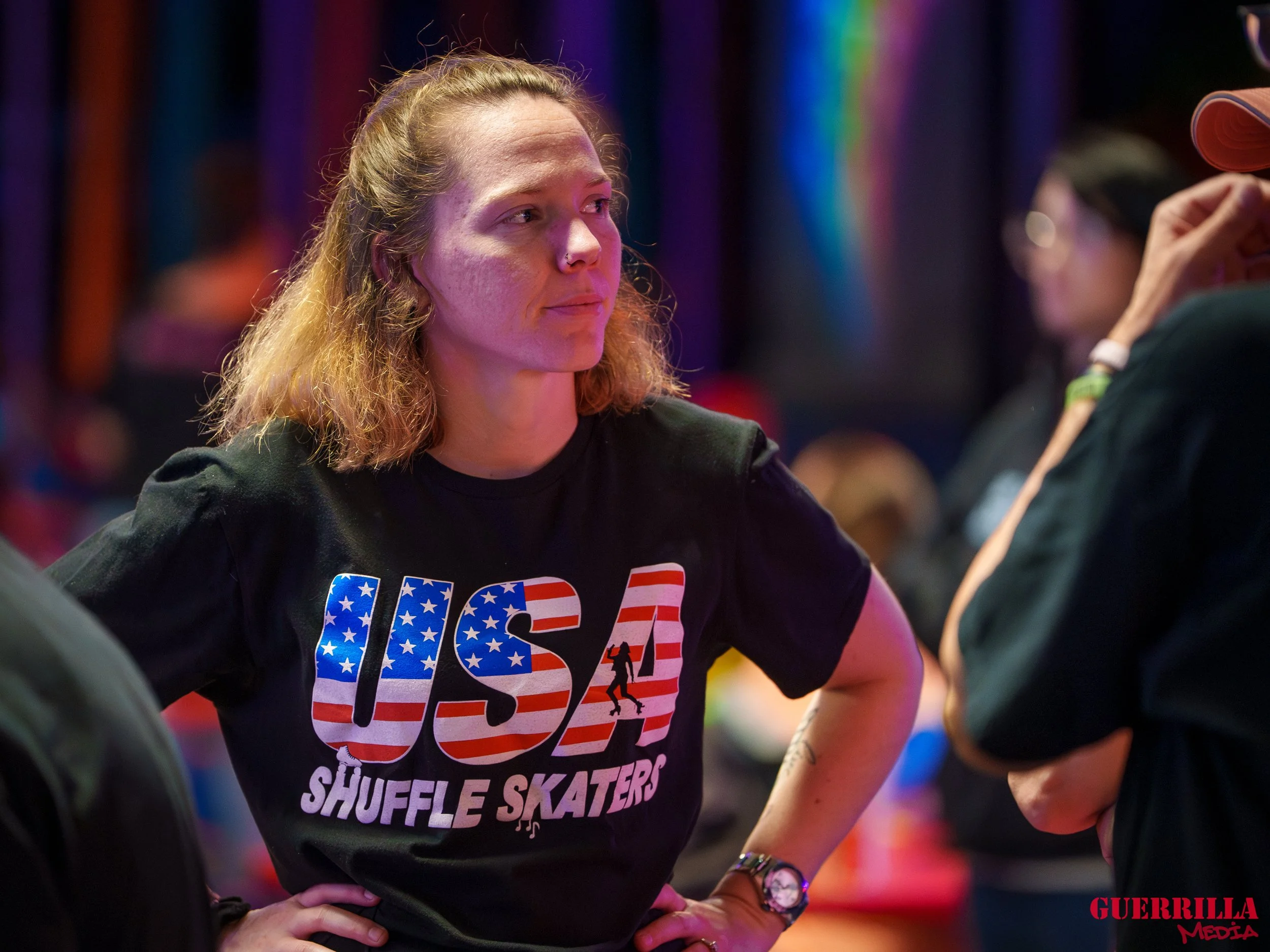 A woman with shoulder-length curly blonde hair, wearing a black T-shirt with an American flag design and the words "US Shuffle Skaters," stands with her hands on her hips, listening to someone off-camera at an indoor skate event with colorful lightin