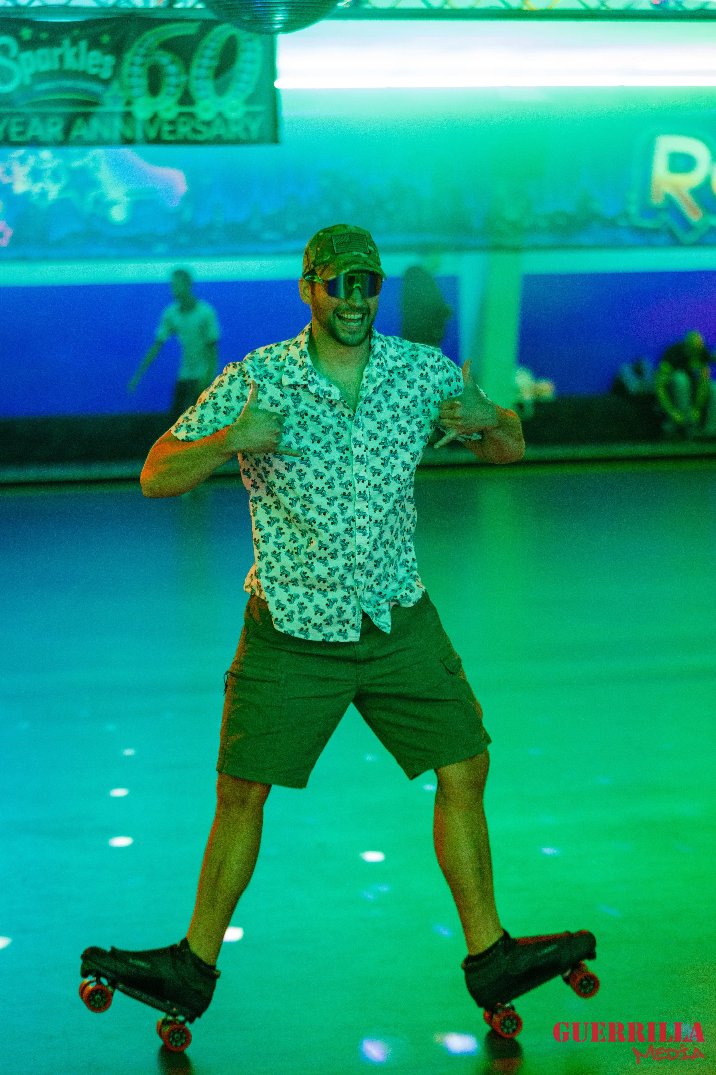A man wearing sunglasses, an animal print shirt, and shorts is roller skating indoors under green lighting, smiling and making a gesture with both hands. A banner in the background marks a 60th anniversary celebration of Sparkles.