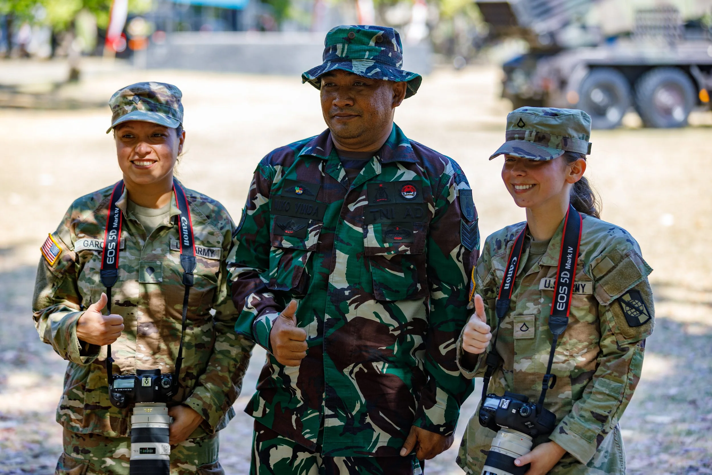 Three people in military camouflage uniforms giving thumbs up, standing outdoors. The person in the middle is a man, and the two women on either side are holding cameras with long zoom lenses. All are smiling and wearing hats.