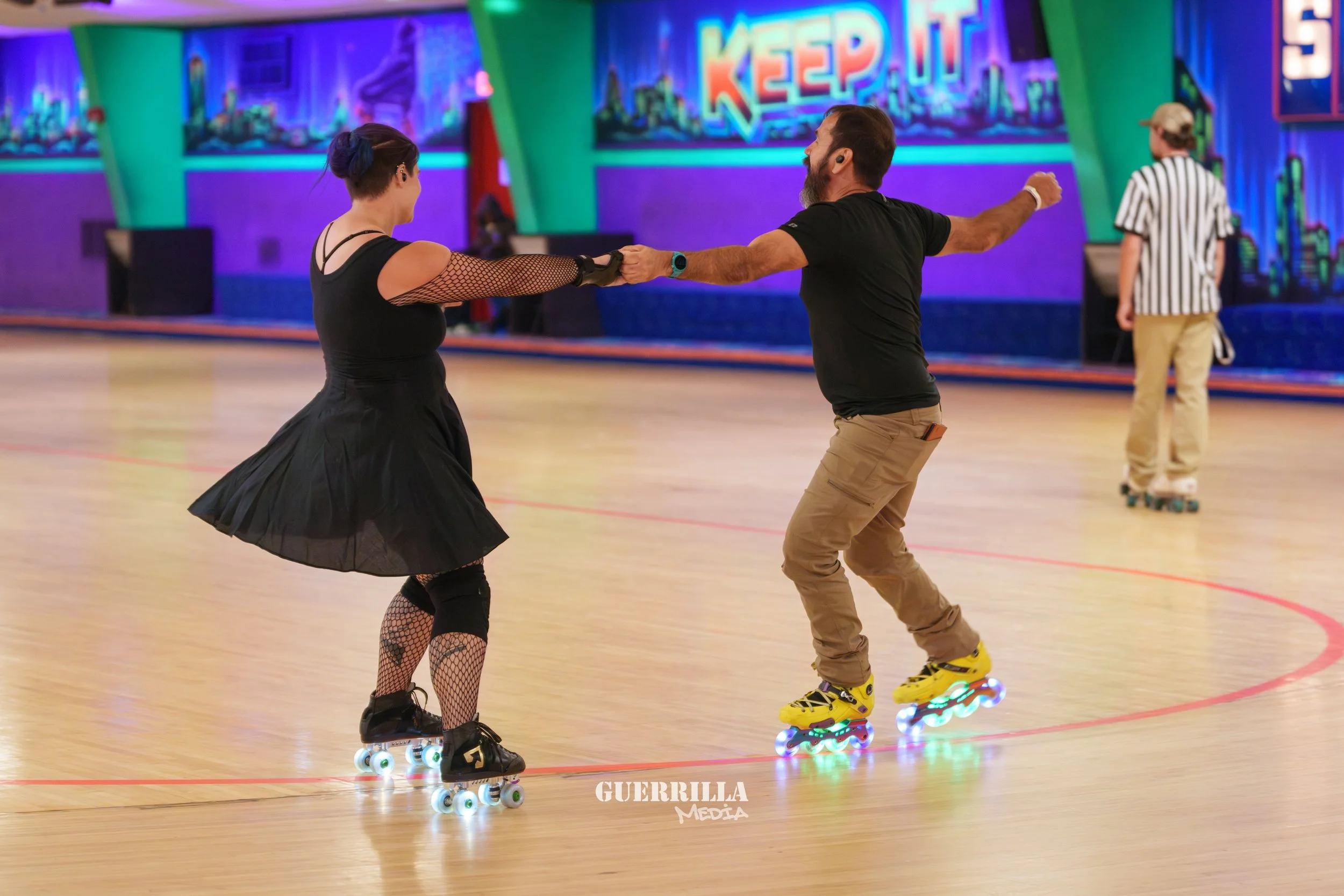 A man and woman roller skating together in an indoor roller rink, holding hands and smiling. The woman is wearing a black dress and fishnet stockings, while the man is dressed in a black t-shirt and khaki pants. There is an official referee in the ba