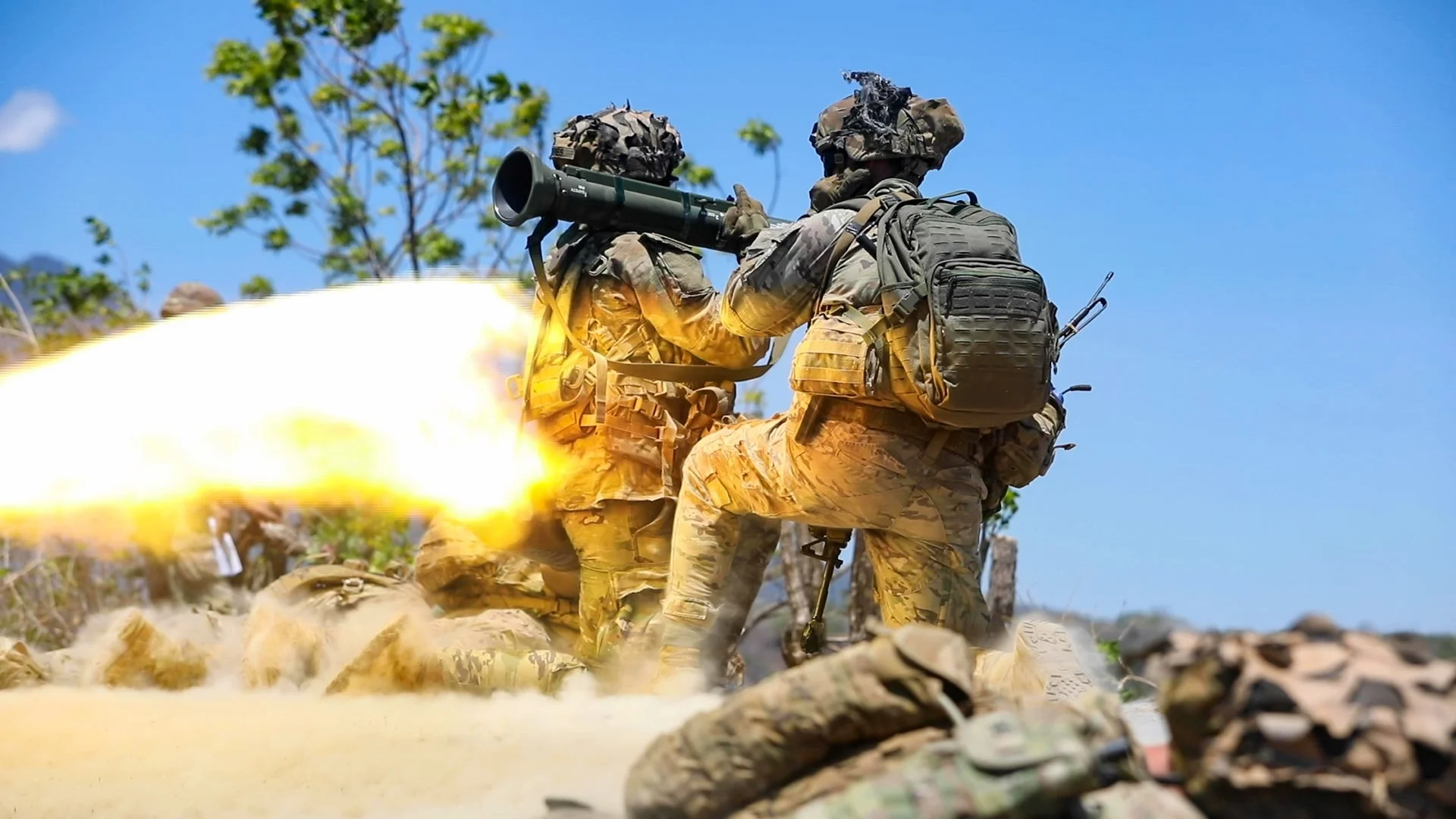 Two soldiers in camouflage uniforms and gear firing a rocket launcher outdoors with trees and a blue sky in the background.
