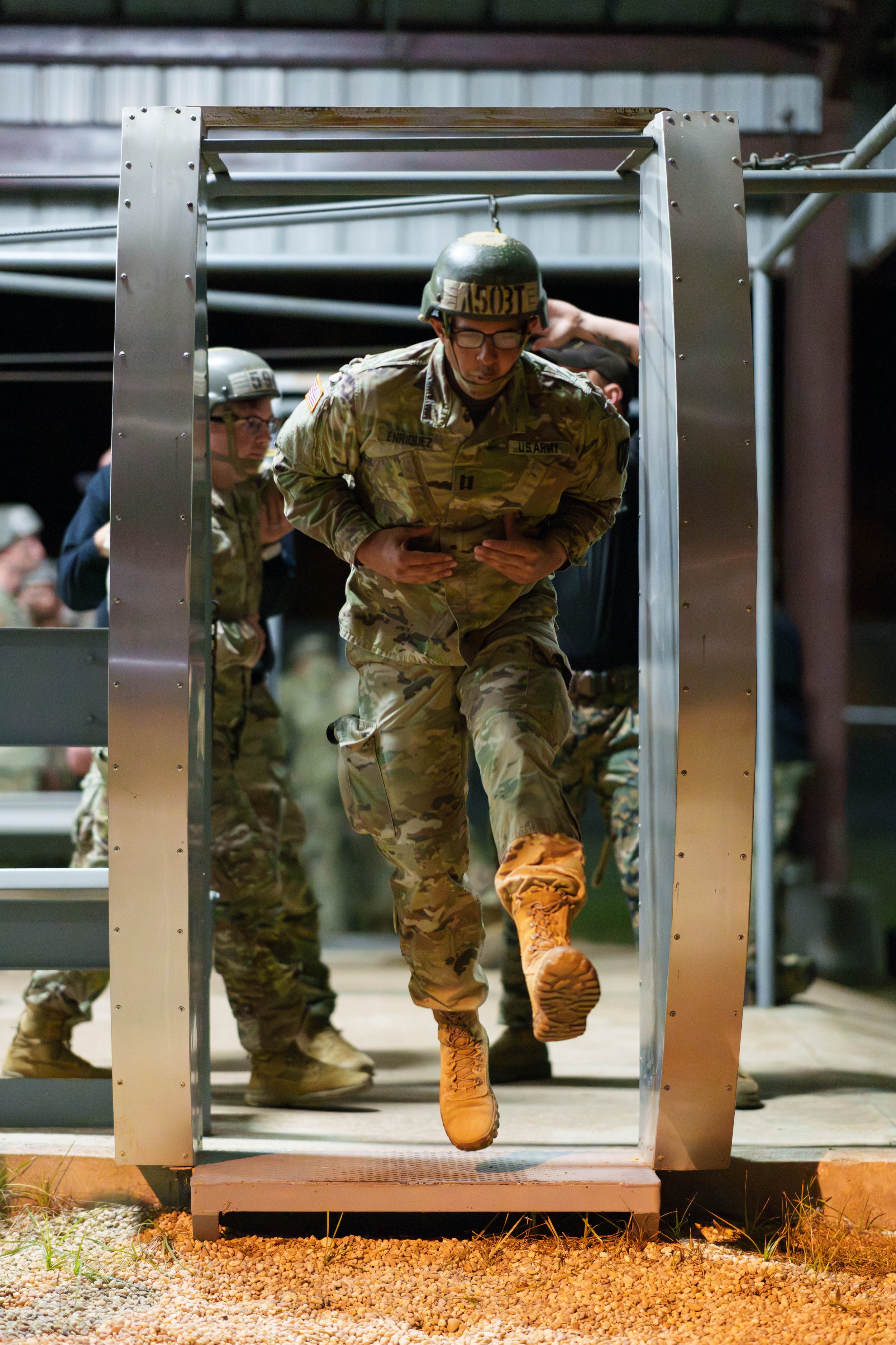 Soldiers in camouflage uniforms and helmets participating in a nighttime military training exercise, running through a metal frame obstacle.