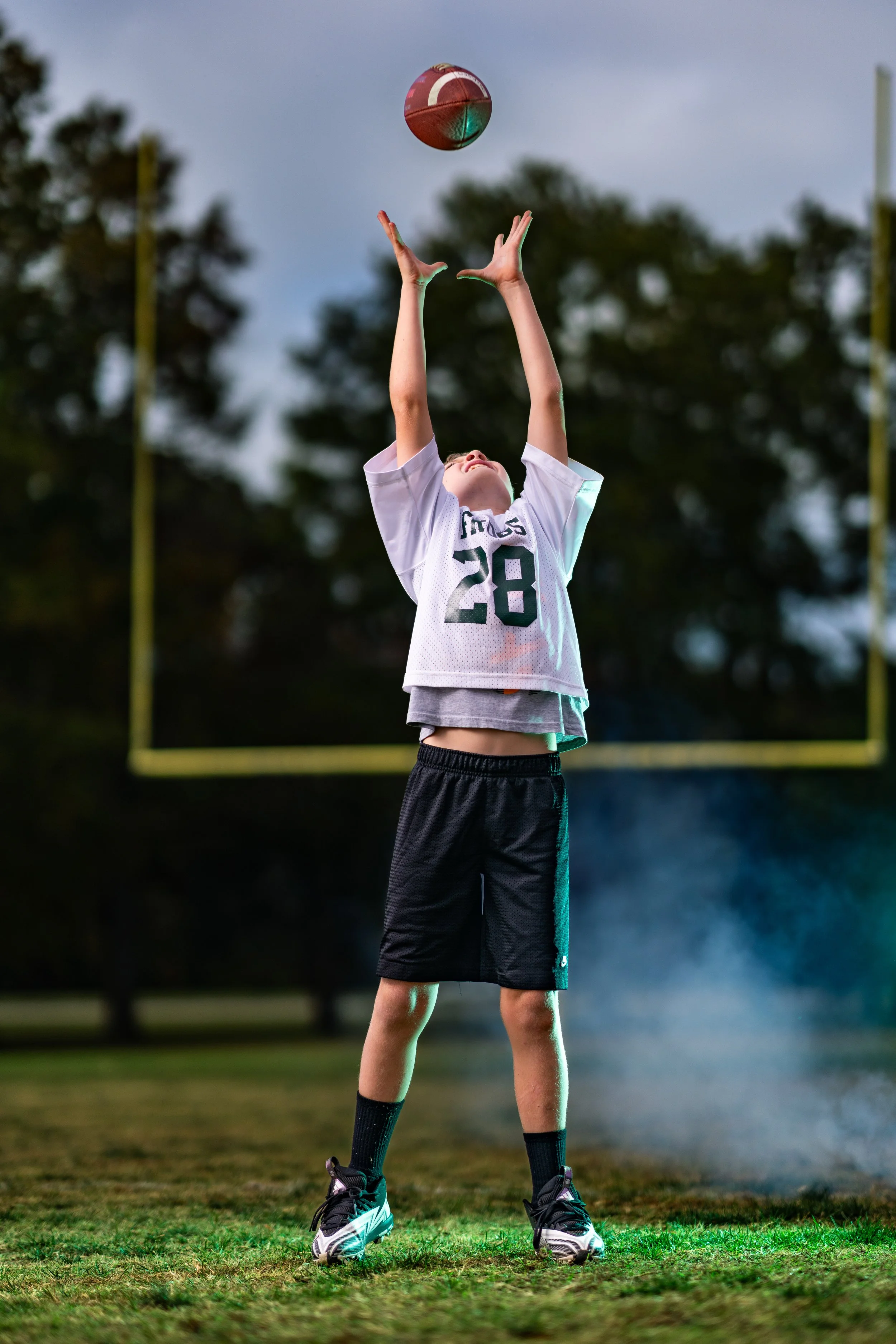 A young football player in a white jersey with the number 28 throws a football on a field during twilight.