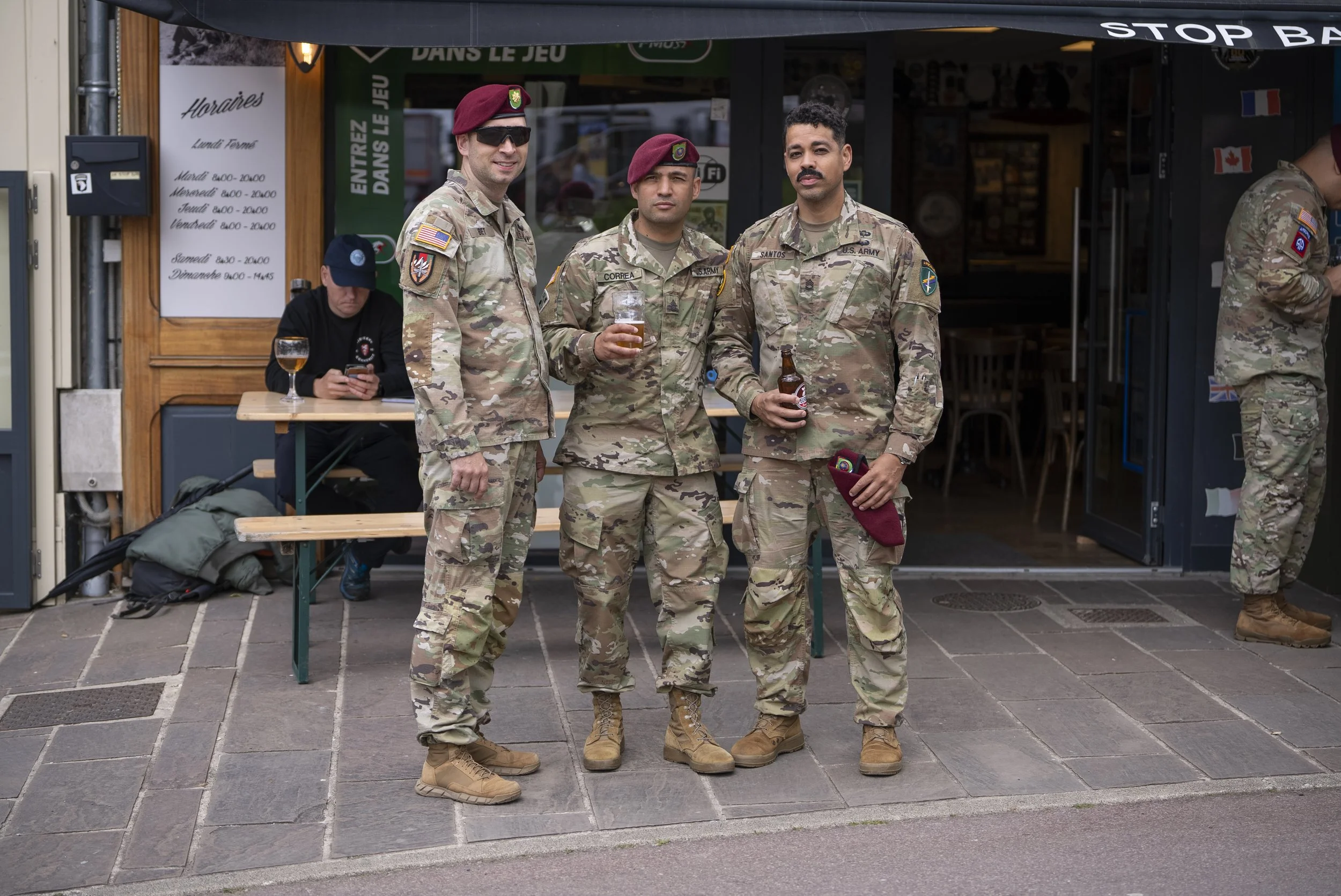 Three soldiers in military camouflage uniforms and berets standing outside a café, holding drinks and posing for a photo. A man is sitting at a table behind them, looking at his phone.