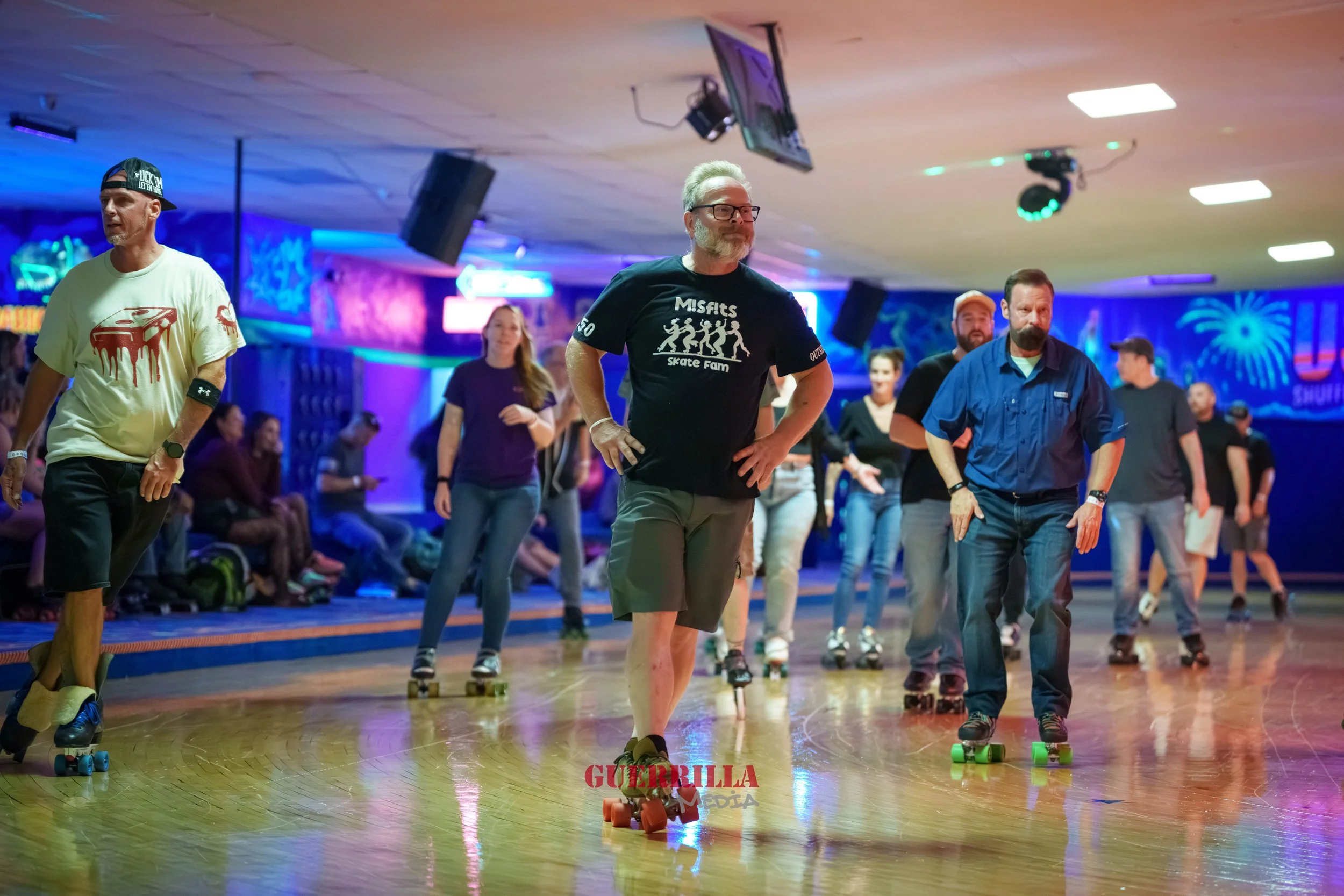 People roller skating in a dimly lit indoor rink with colorful neon lights and a mural in the background.