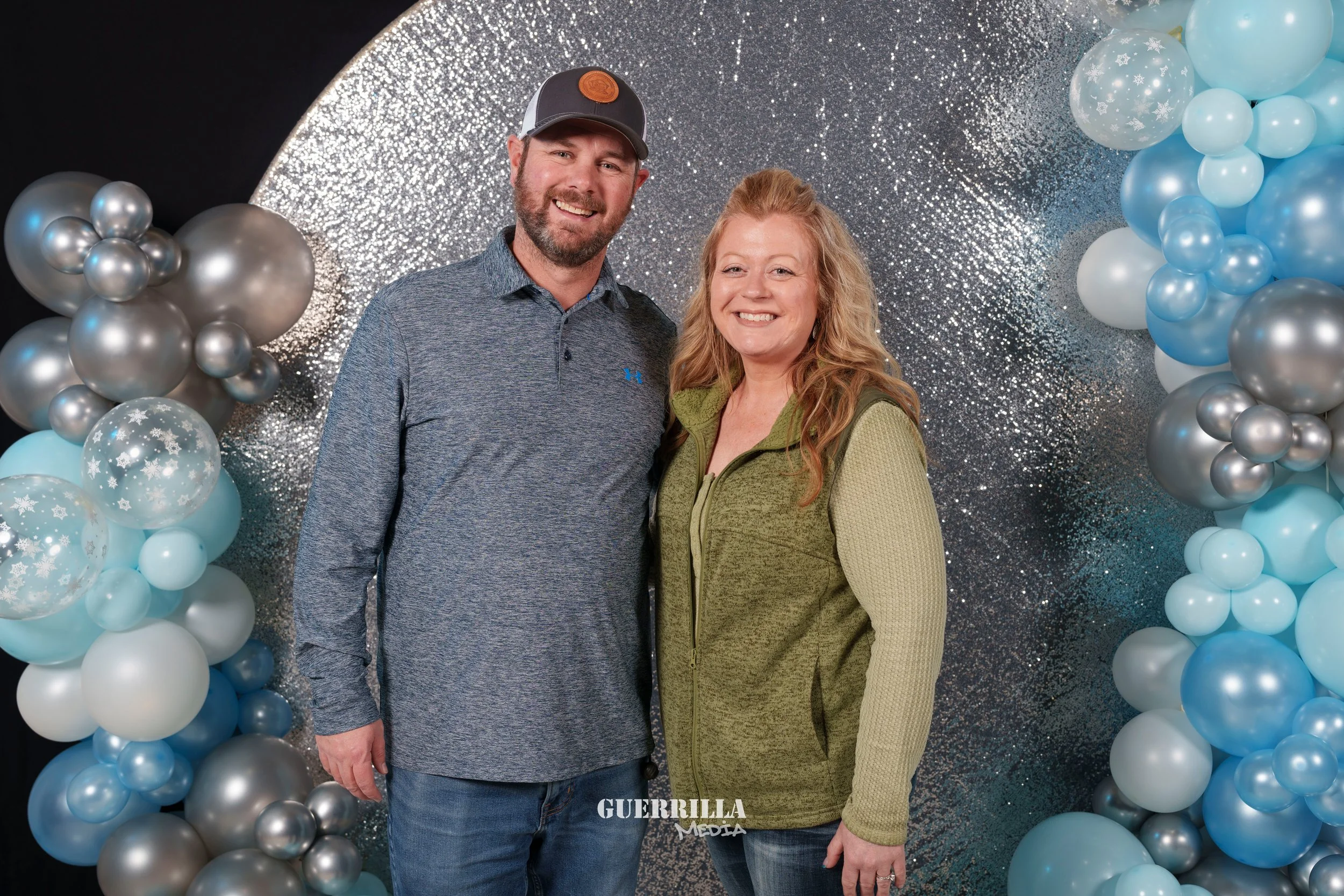 A man and woman standing together smiling in front of a festive backdrop with blue, silver, and white balloons and a glittery background.