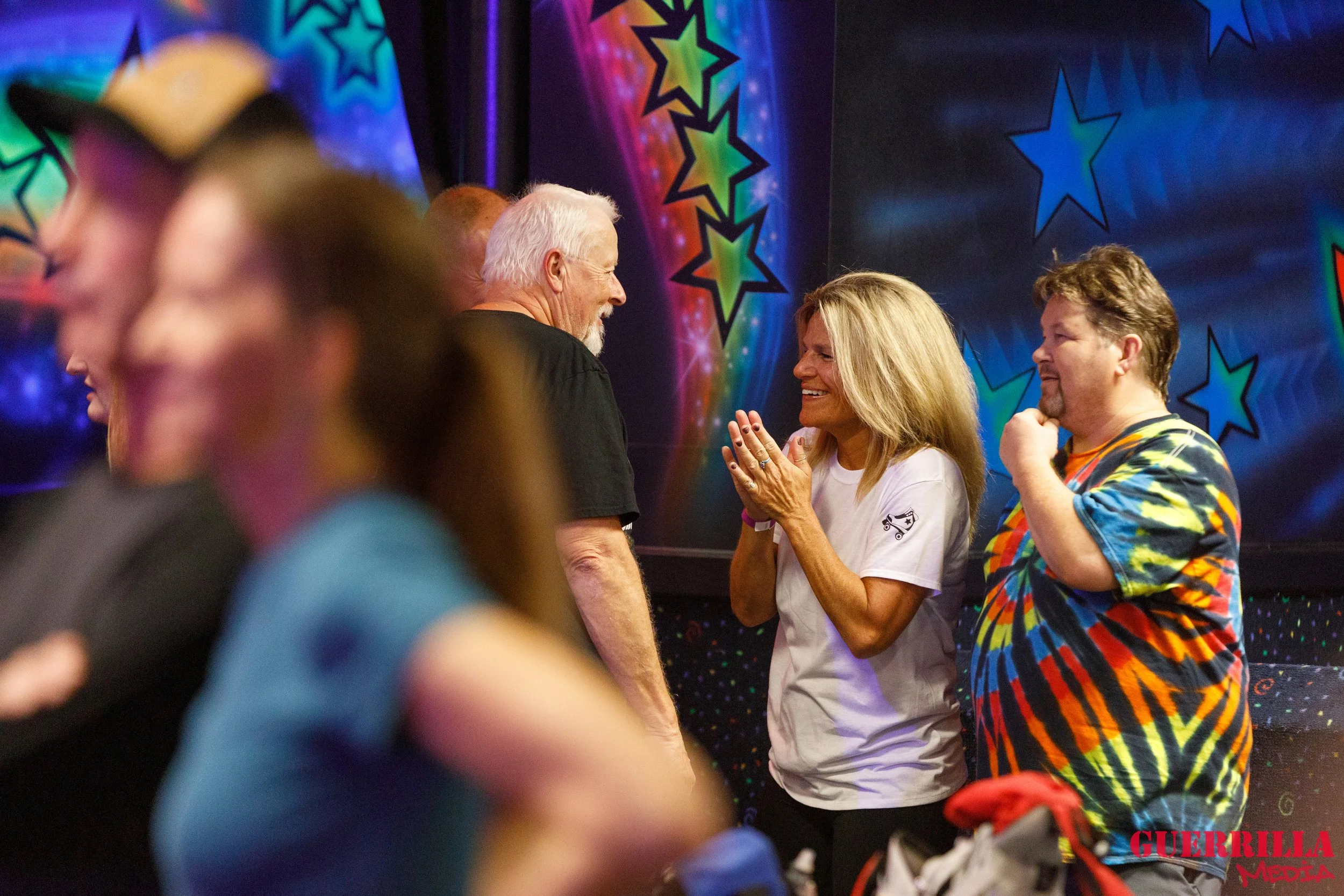 A group of people at a party or gathering are smiling and talking, with a colorful star-themed backdrop in the background. The focus is on a woman in a white T-shirt with short blonde hair, smiling and engaging with an older man with white hair and a