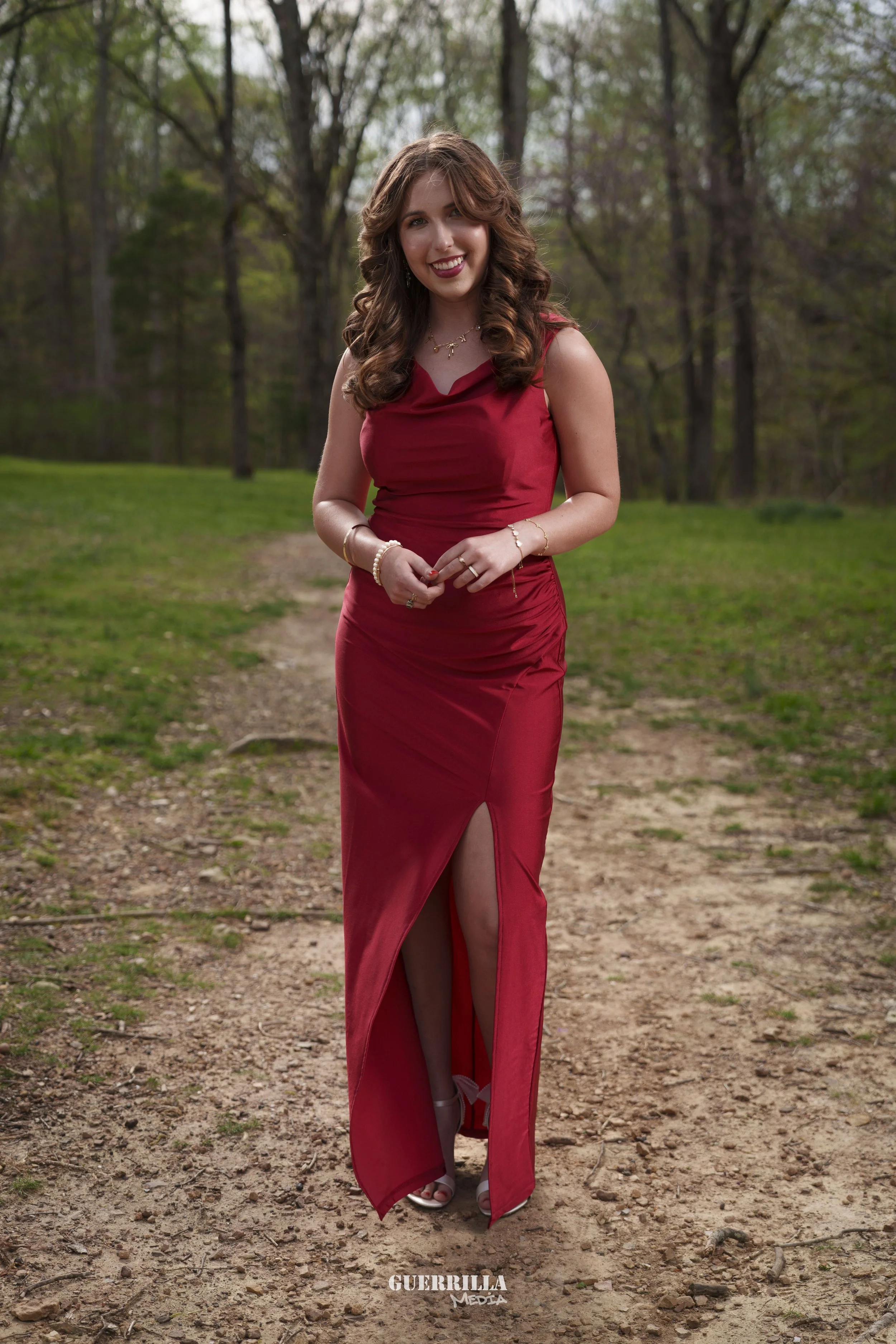 A young woman in a red satin evening gown with a slit walking on a dirt path in a wooded park.