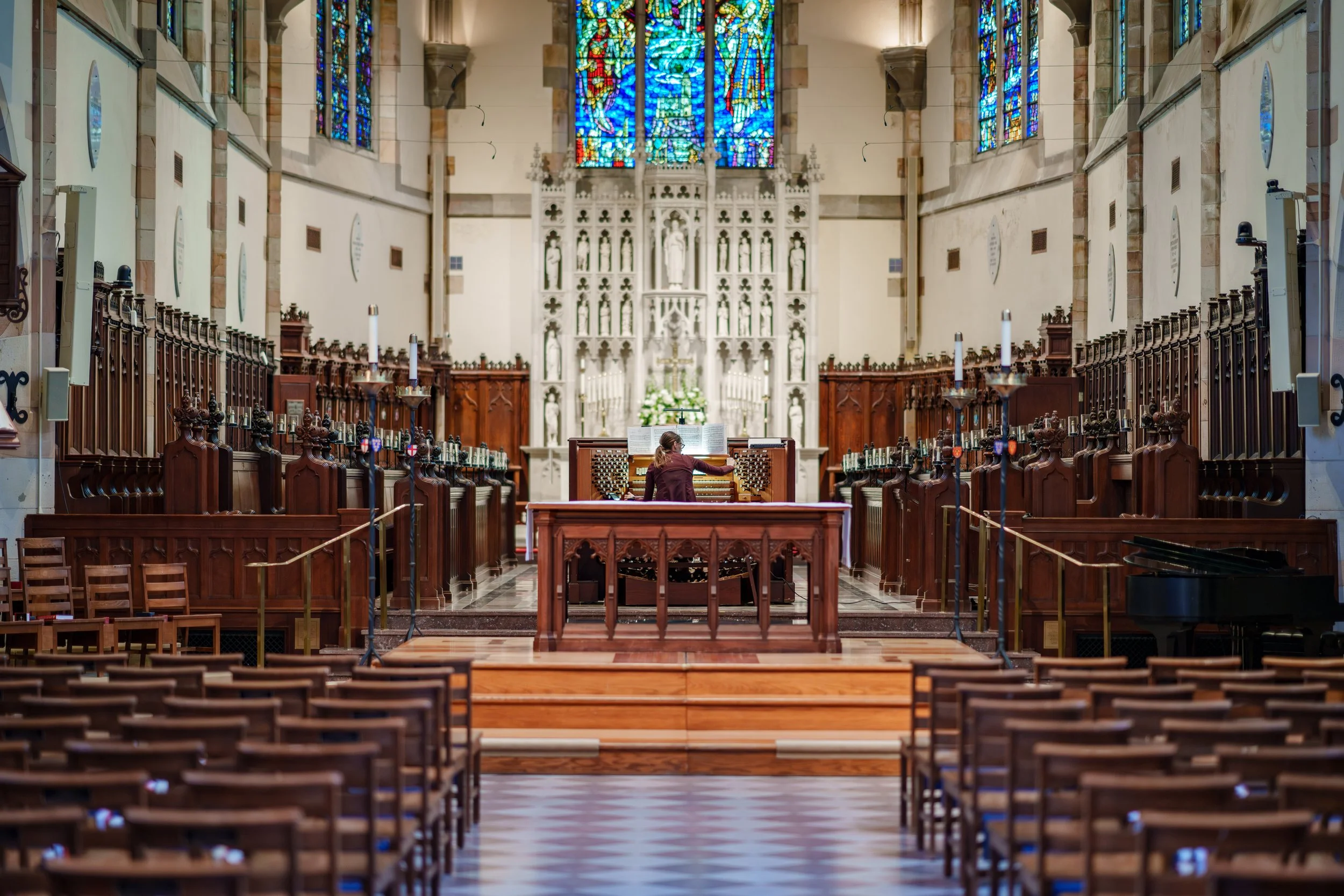 Interior of a church with a woman playing a pipe organ at the front, wooden pews, stained glass windows, and an altar in the background.