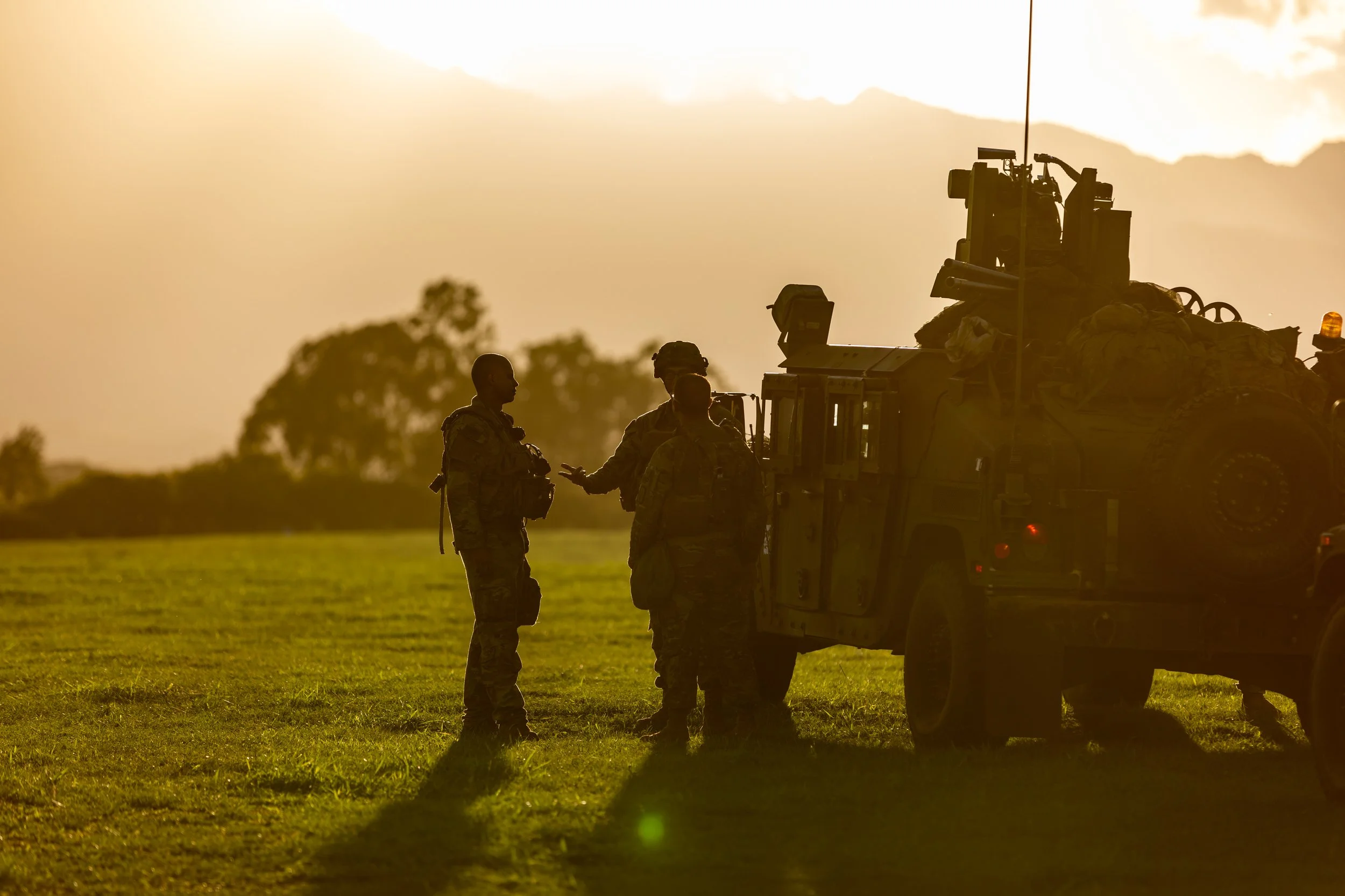Silhouettes of three soldiers in uniform having a conversation next to military vehicle during sunset in an open field.
