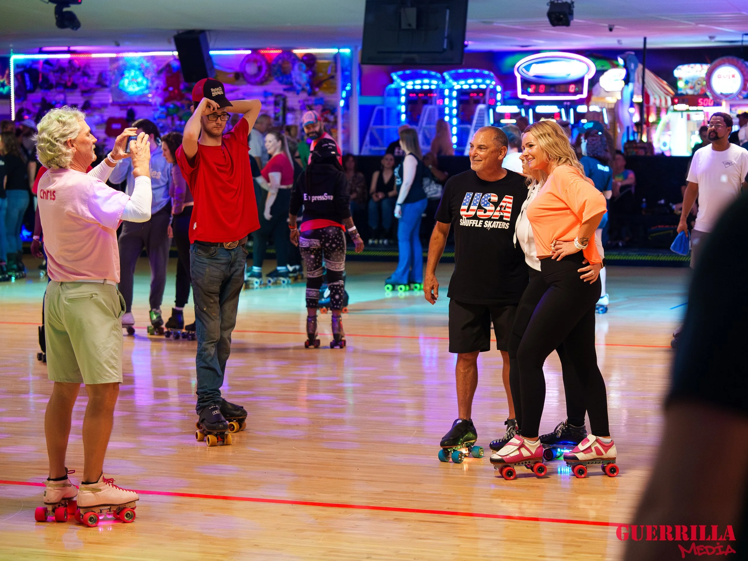 People roller skating at an indoor rink with neon lights and arcade games in the background. Some are taking pictures and socializing, enjoying a fun, lively atmosphere.
