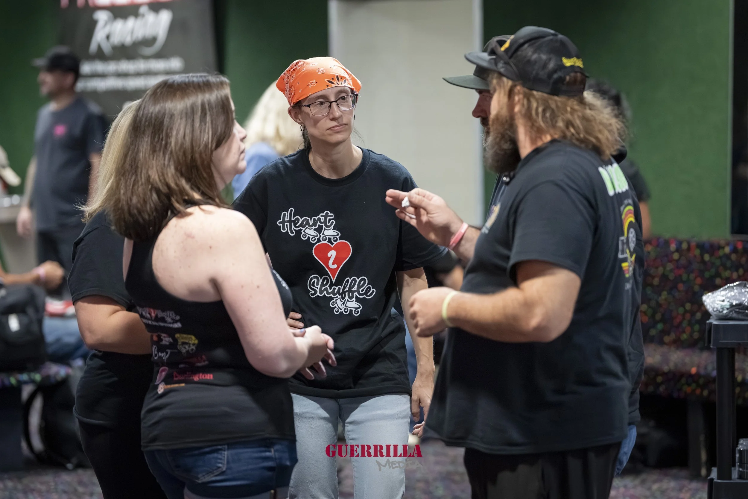 Four people engaged in a conversation at an indoor event. The woman in the center is wearing glasses and an orange bandana, looking seriously at the man in the black cap and beard who is speaking. The woman on the left has shoulder-length brown hair 