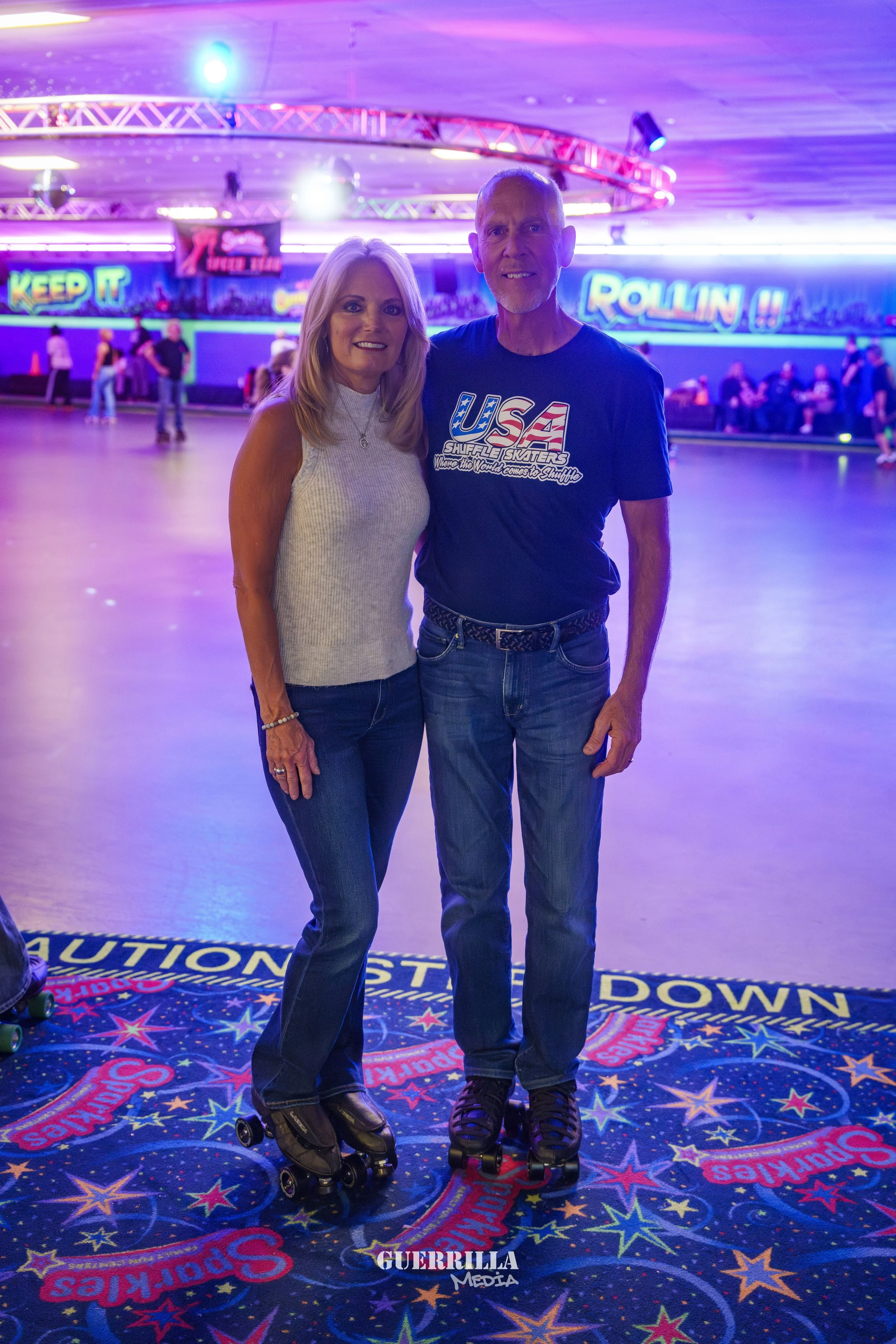 A smiling man and woman stand on the roller rink floor with roller skates, surrounded by neon lights and colorful signs in the background.