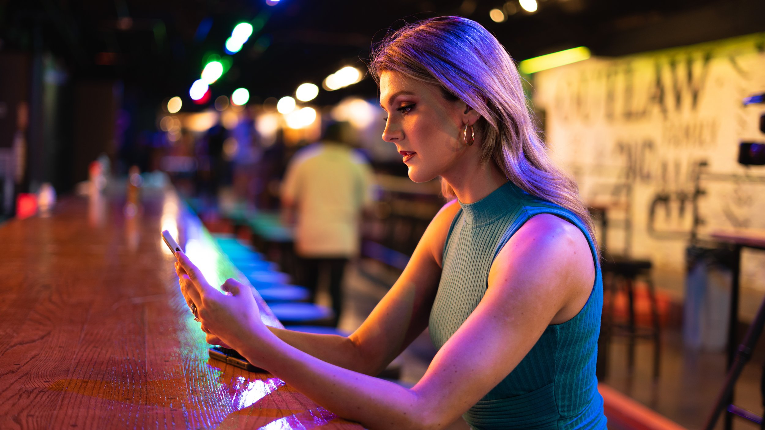 Woman with blonde hair and hoop earrings sitting at a bar, looking at her phone, with colorful neon lights in background.
