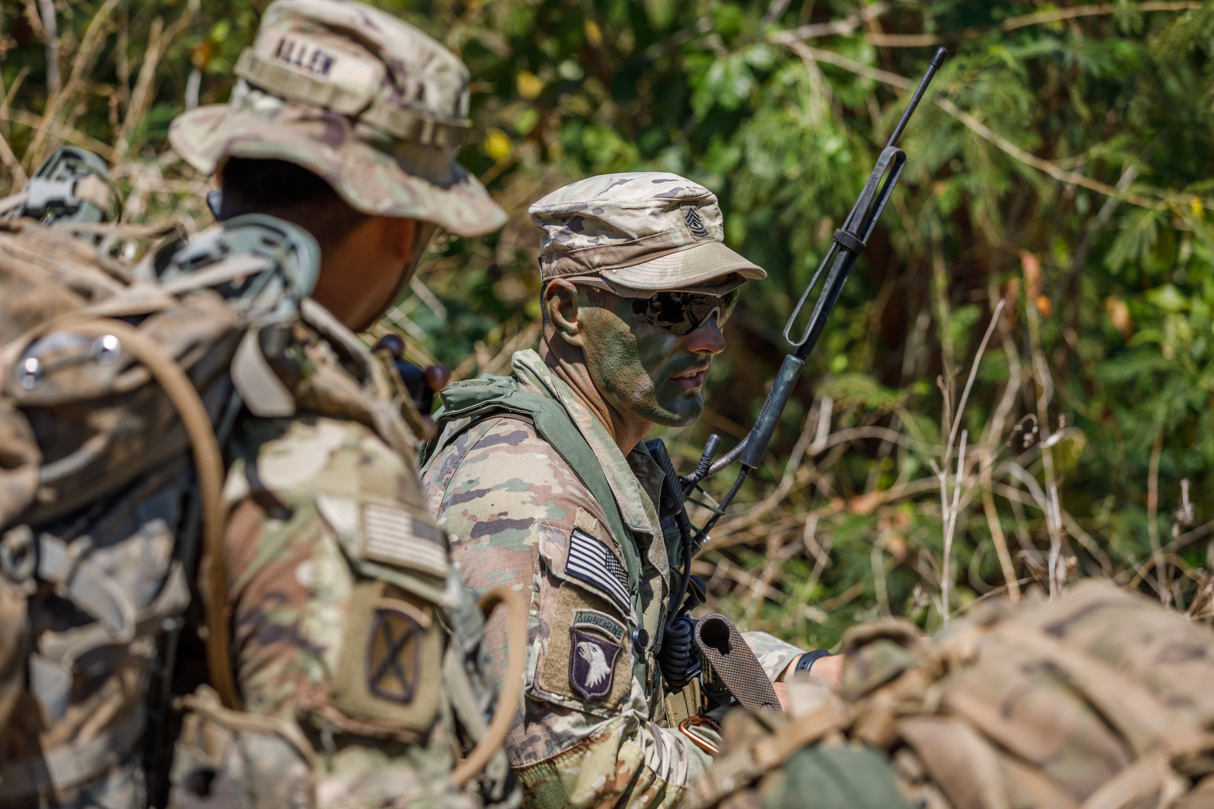 Two soldiers in camouflage uniforms and gear, with one wearing sunglasses and holding a radio antenna, sitting in a forest.