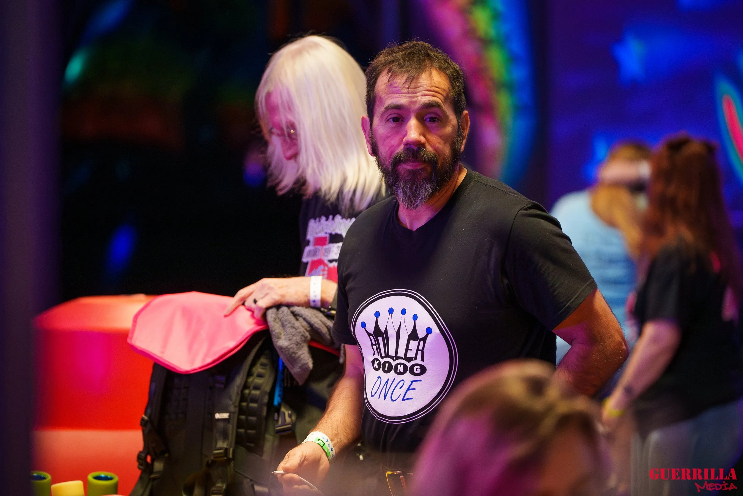 Man in black t-shirt with 'Killer Kong Once' logo at roller skating event, surrounded by people, with colorful neon lights in background.