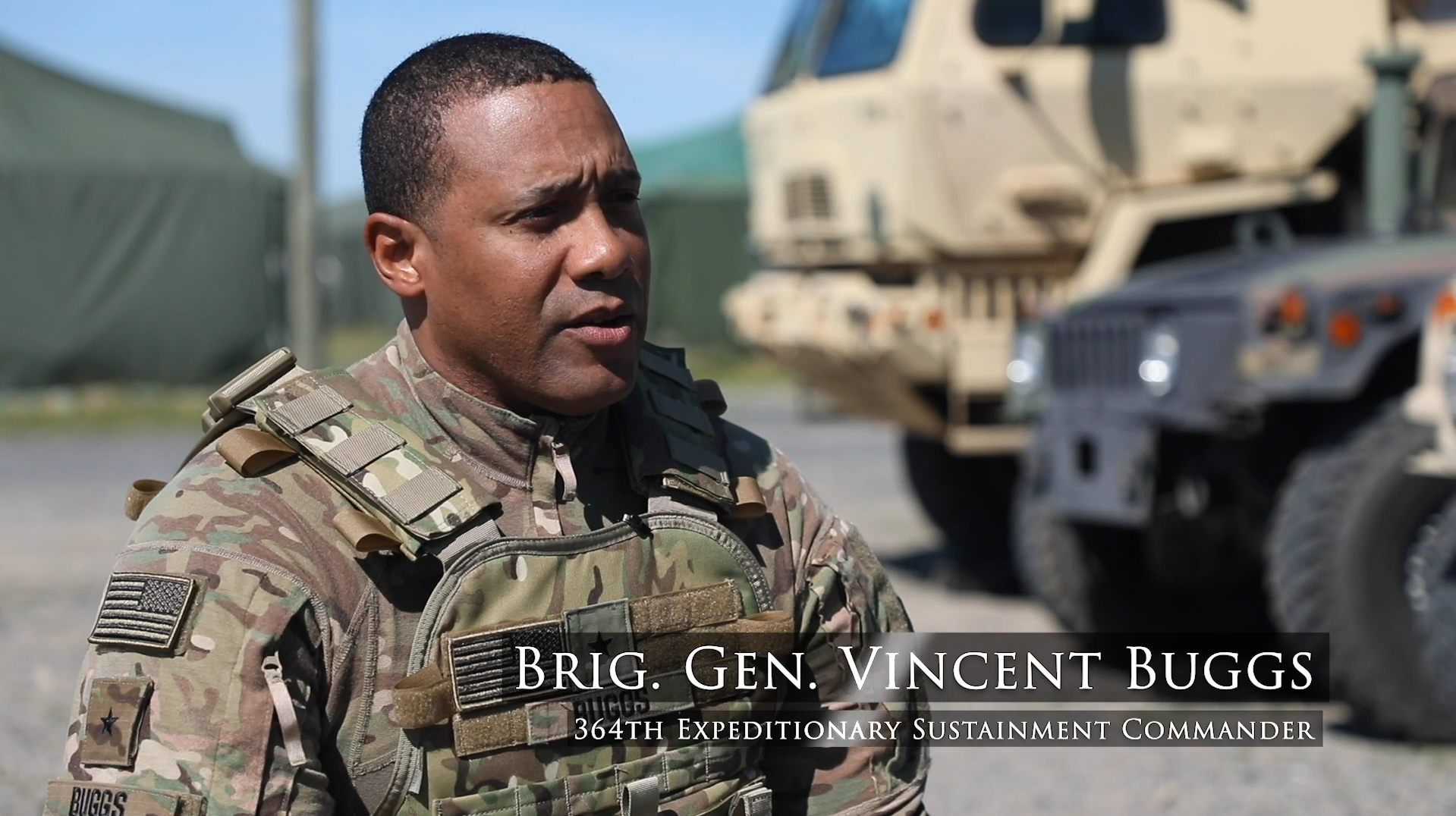 A military officer in camouflage uniform with a name tag that says 'BUGGS', speaking outdoors with military vehicles in the background. The text overlay reads 'BRIG. GEN. VINCENT BUGGS, 364TH EXPEDITIONARY SUSTAINMENT COMMAND.'