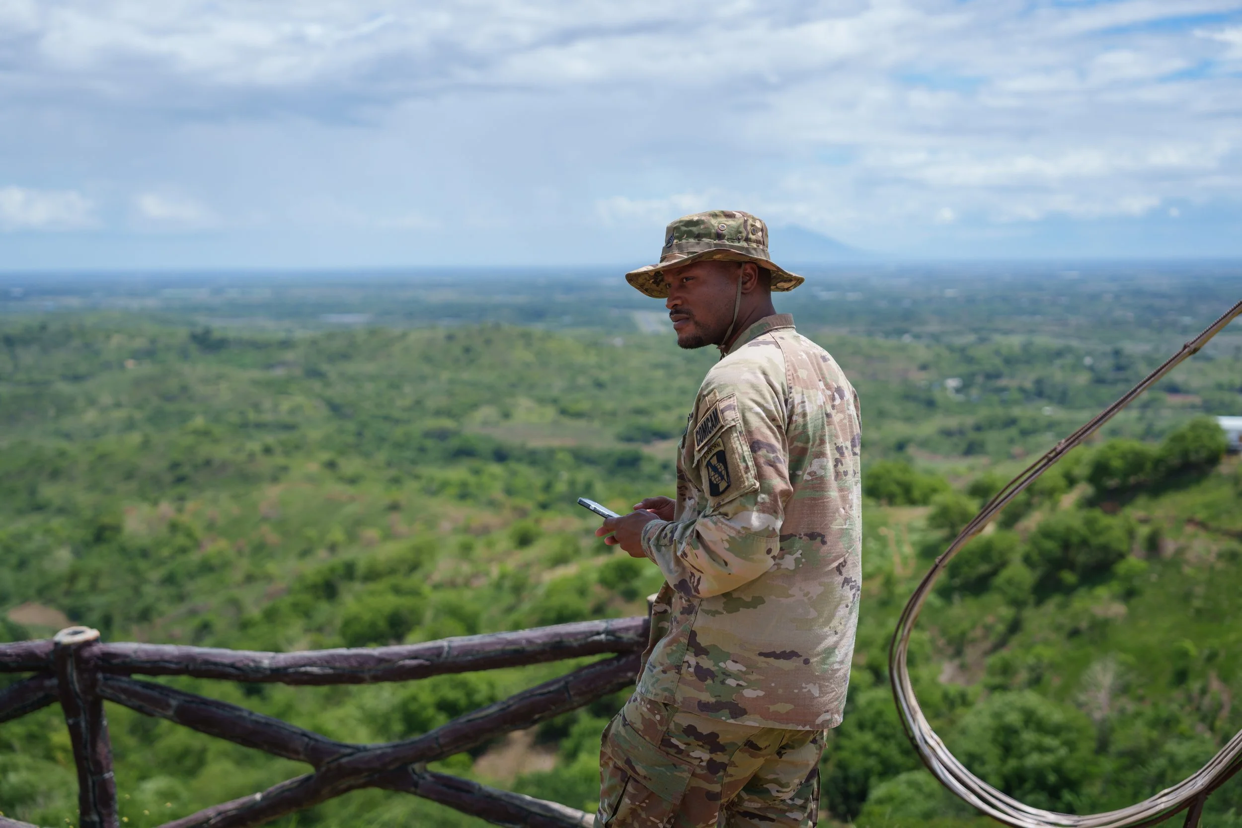 A soldier in camouflage uniform standing on a wooden lookout platform in a lush green landscape, looking at a mobile device.