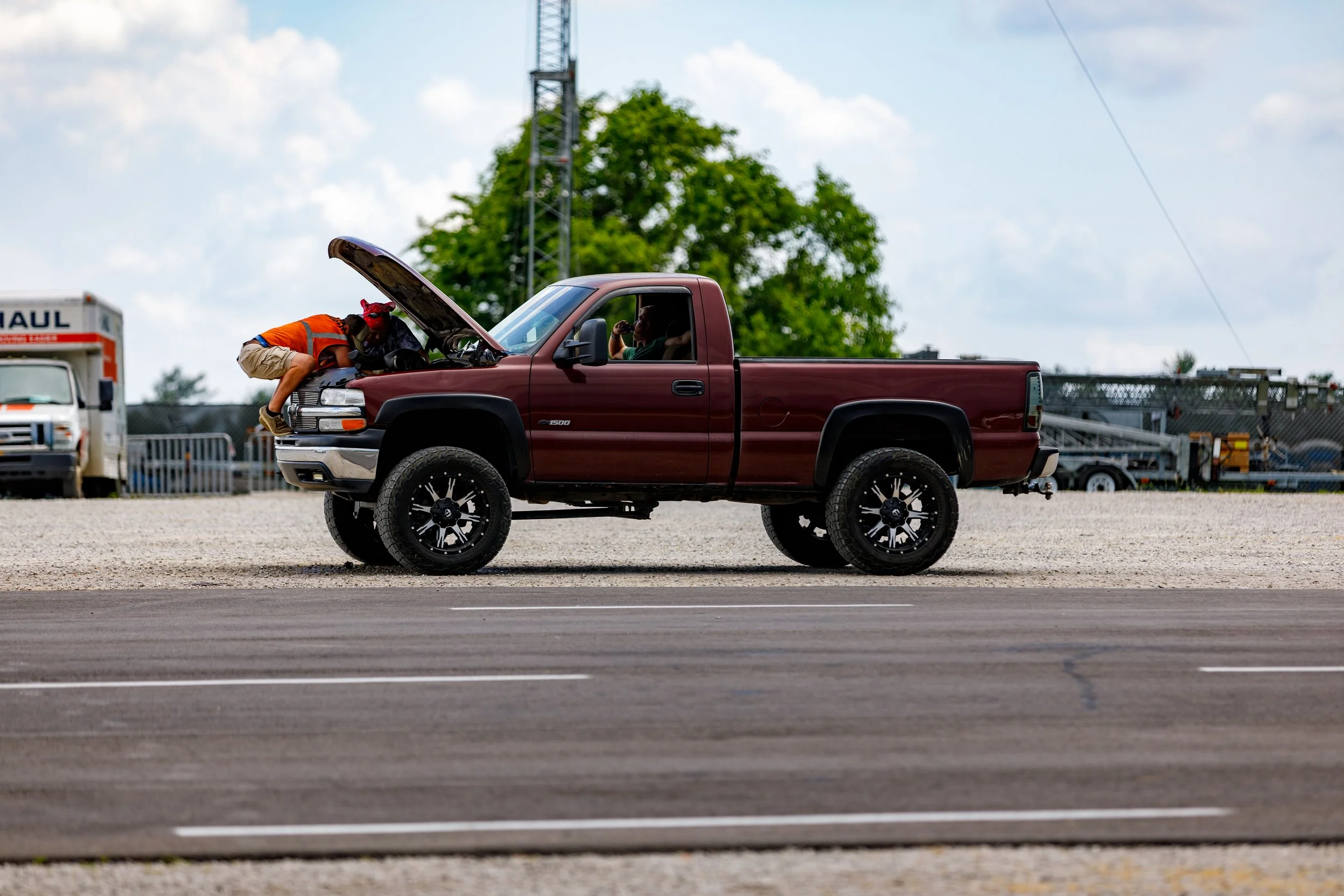 A person leaning over the open hood of a maroon pickup truck, with another person sitting inside, on a gravel parking lot during the day.