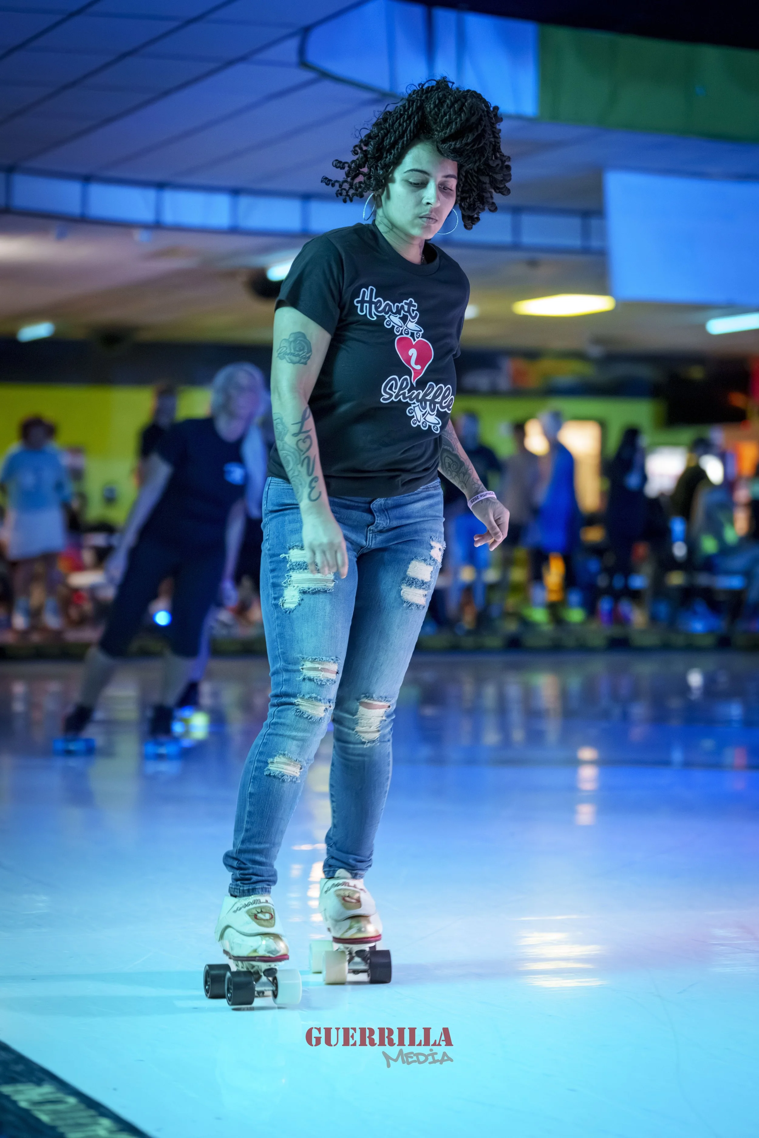 A woman roller skating indoors at a roller rink, wearing a black t-shirt with the text 'Heart & Shuffle,' distressed jeans, and white roller skates. She has curly hair and tattoos on her arms, and there are other skaters in the background.