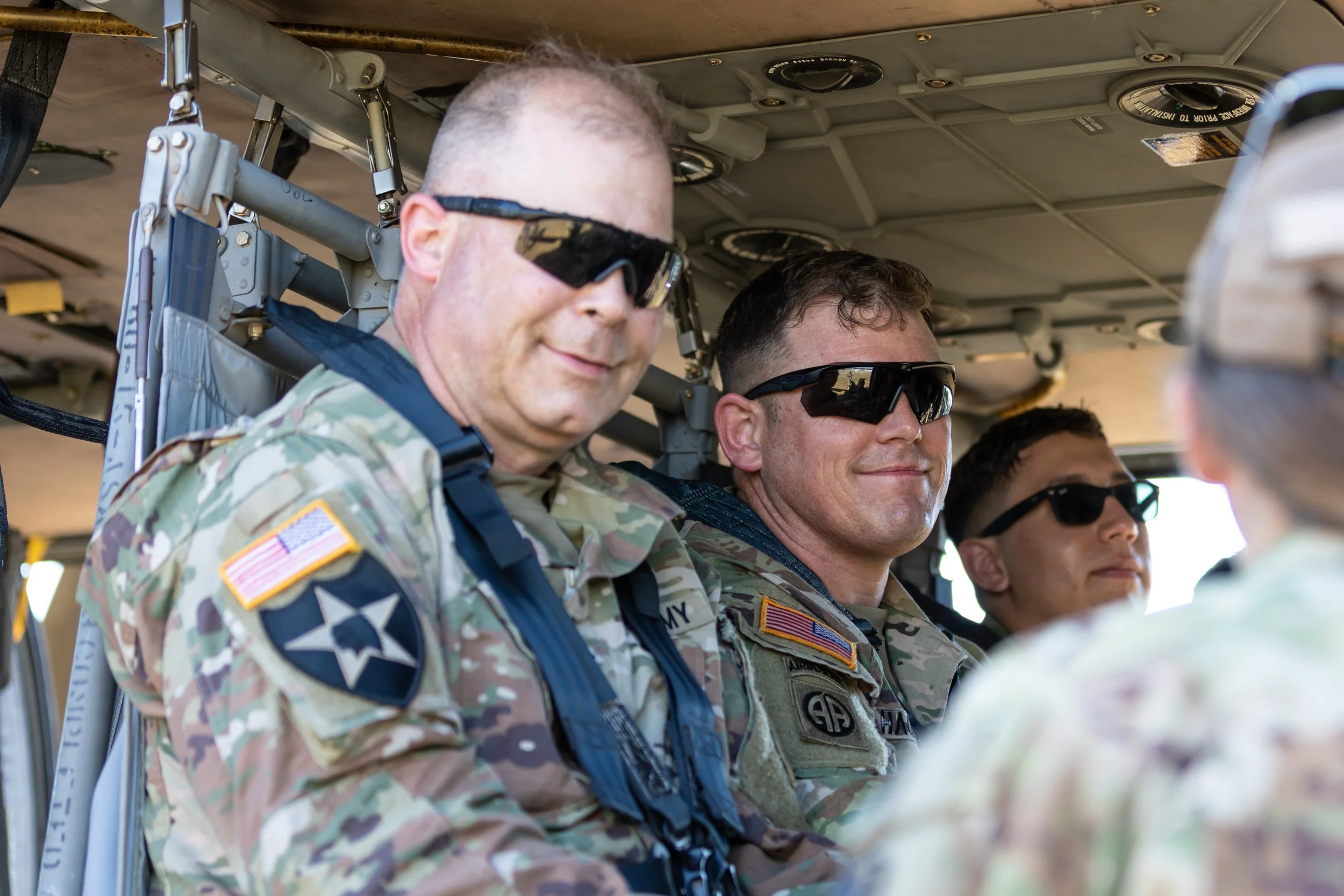 Three military personnel wearing camouflage uniforms and black sunglasses inside a helicopter.