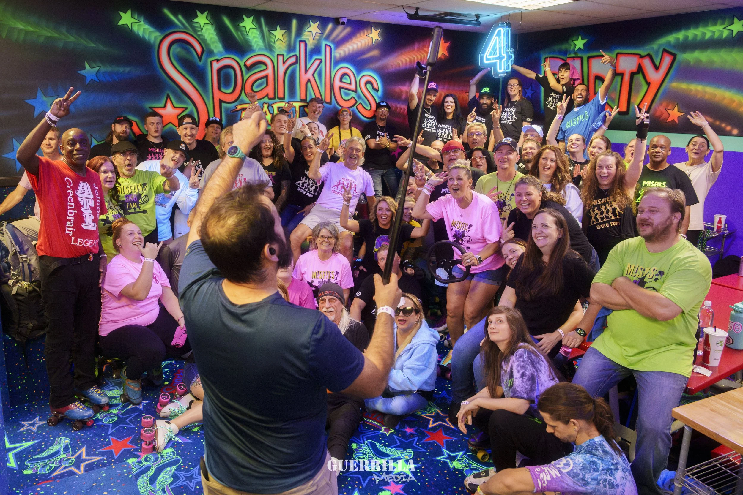 A large group of people celebrating at a roller skating rink with a colorful, neon-lit background that says 'Sparkles Skate Party.' Many are wearing casual and themed clothing, some holding skateboards, and others taking photos, showing excitement an