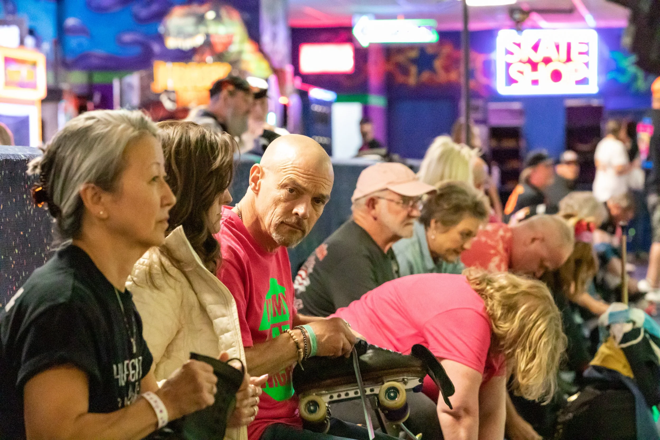 Group of people sitting in a skate shop, some resting or waiting, with neon signs and colorful graffiti art in the background.