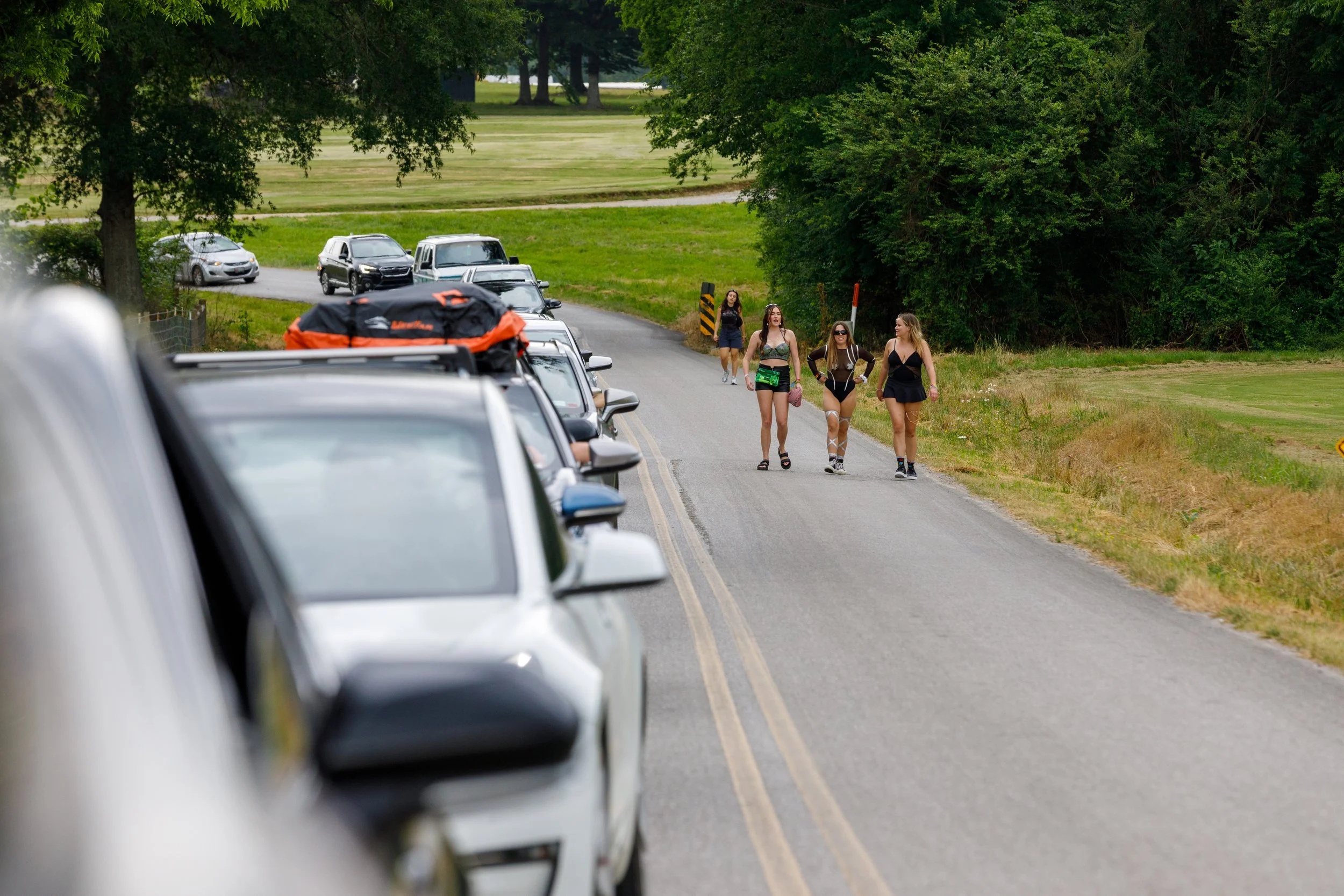 Four women walking on a rural road with parked cars on the side, trees in the background, and some cars in motion.