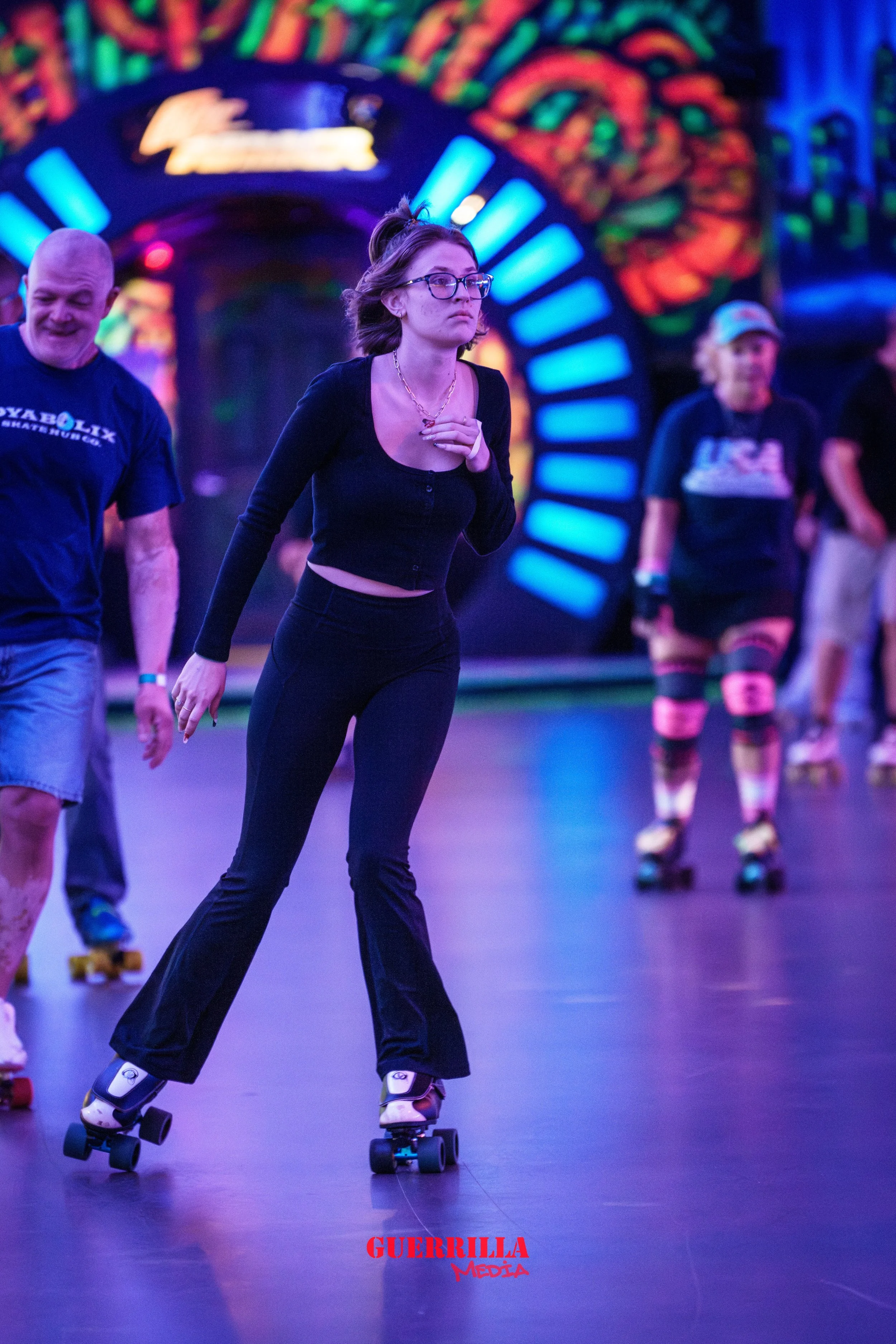 A young woman roller skating indoors with blue and purple lighting, wearing black pants and a black top, with colorful neon abstract artwork in the background.