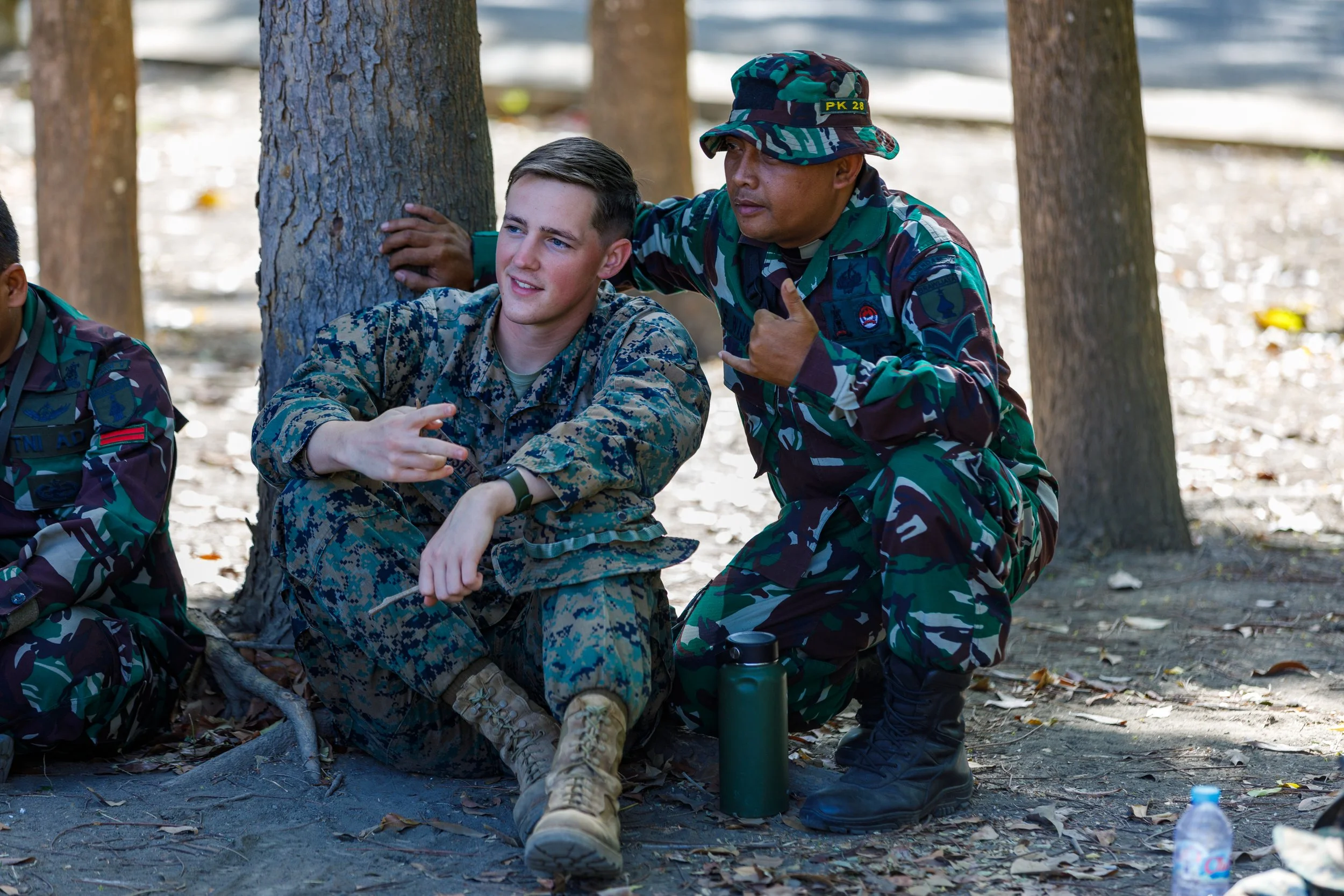 Two military personnel in camouflage uniforms sitting and kneeling on the ground, engaging in conversation in a wooded outdoor setting.