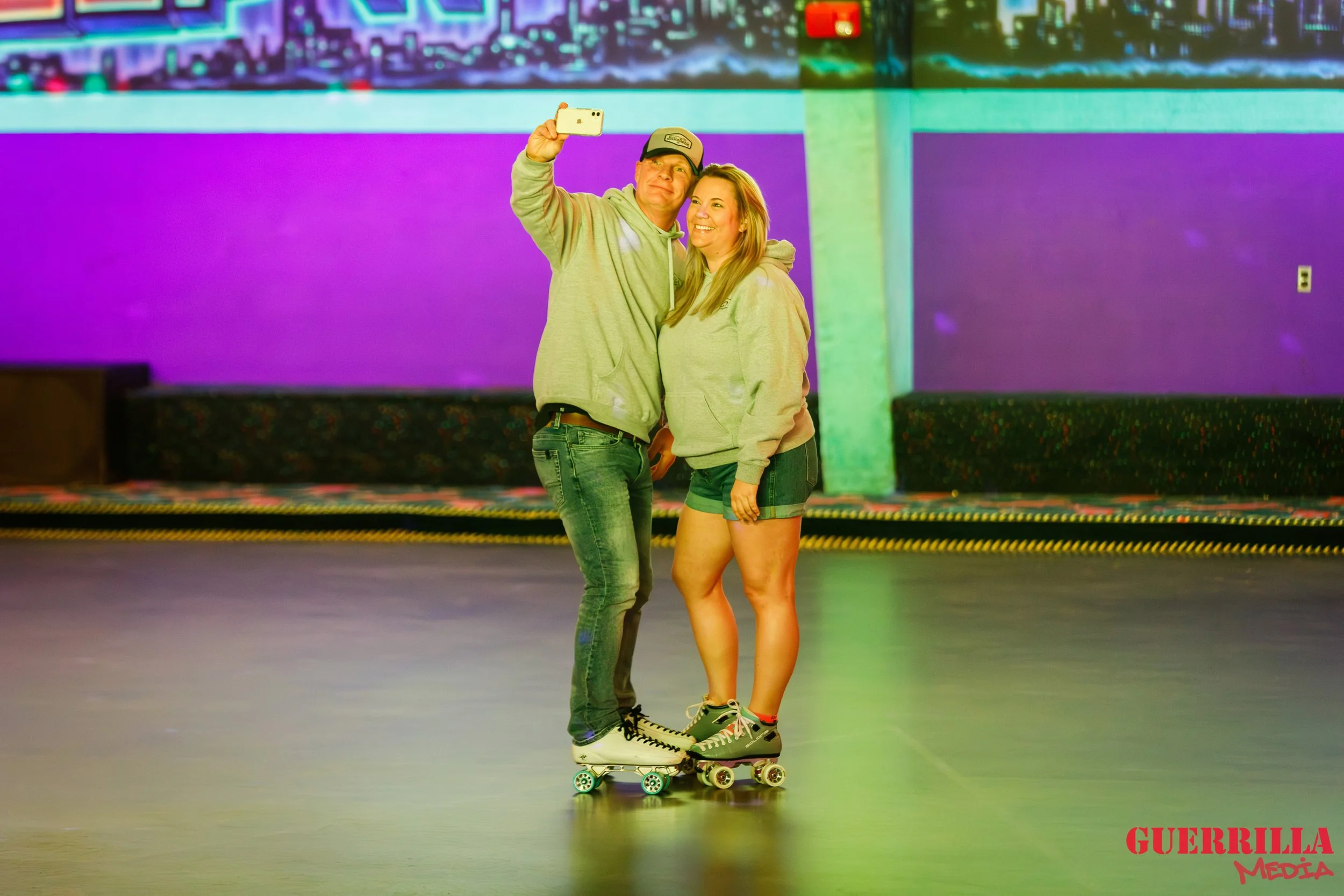 A young man and woman stand close together on roller skates, taking a selfie inside a colorful roller skating rink with purple, green, and blue lighting.