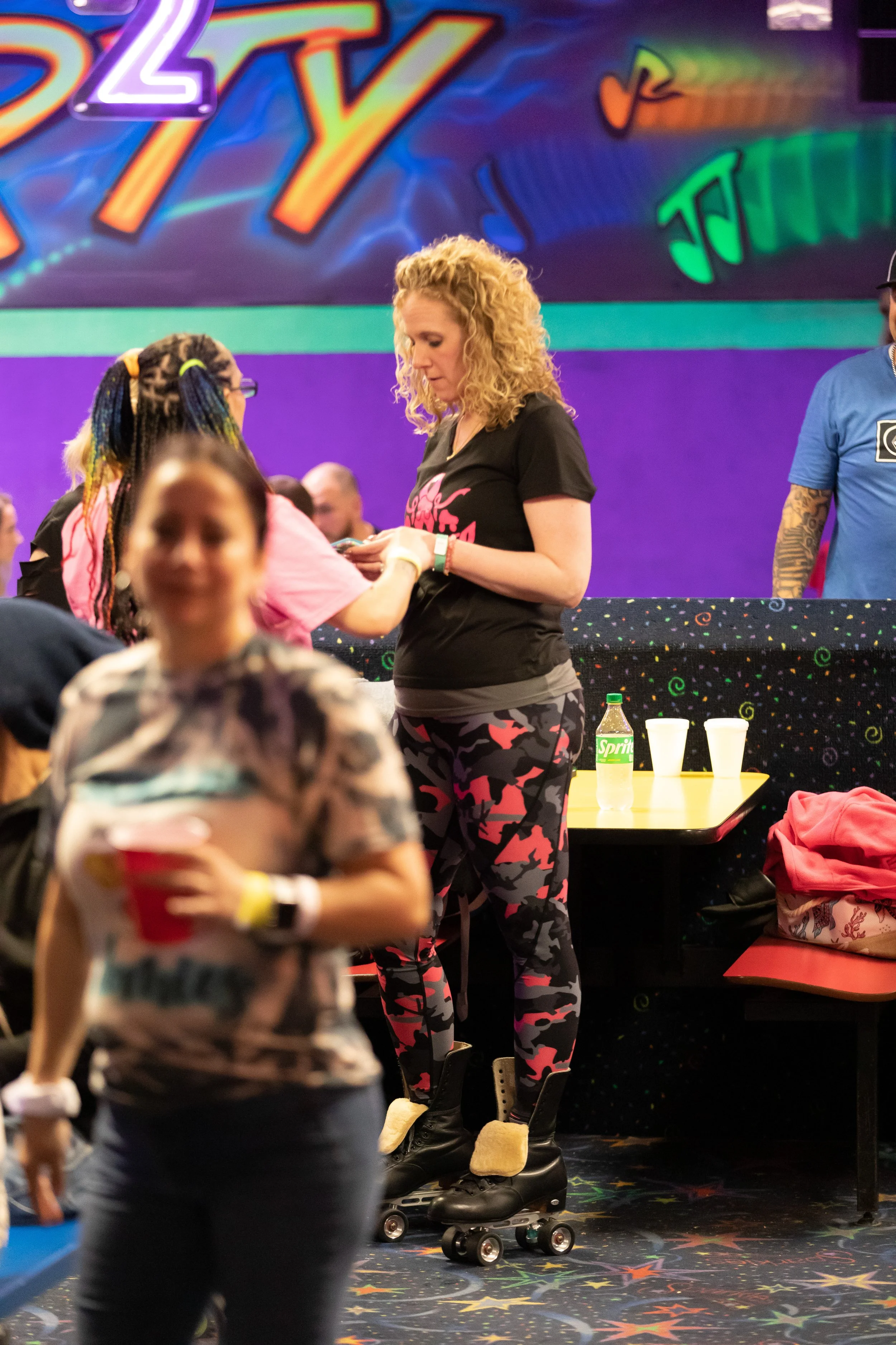 A woman with blonde curly hair standing on roller skates in an entertainment venue, interacting with another person wearing glasses and colorful clothing. In the background, a colorful mural and people at a bar or counter, with beverages on a table.