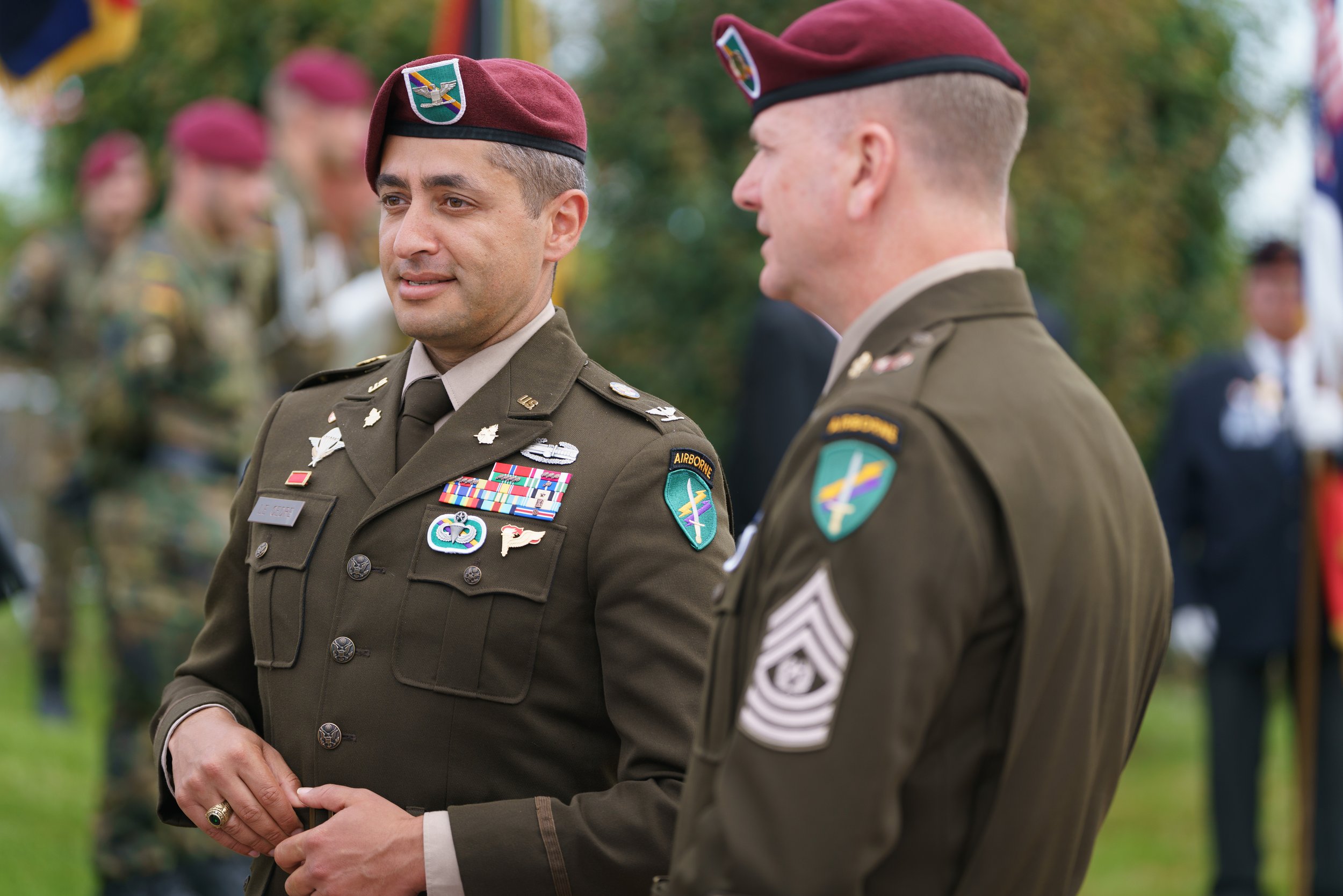 Two military officers in uniform and maroon berets standing outdoors during a ceremony, with other soldiers and people visible in the background.
