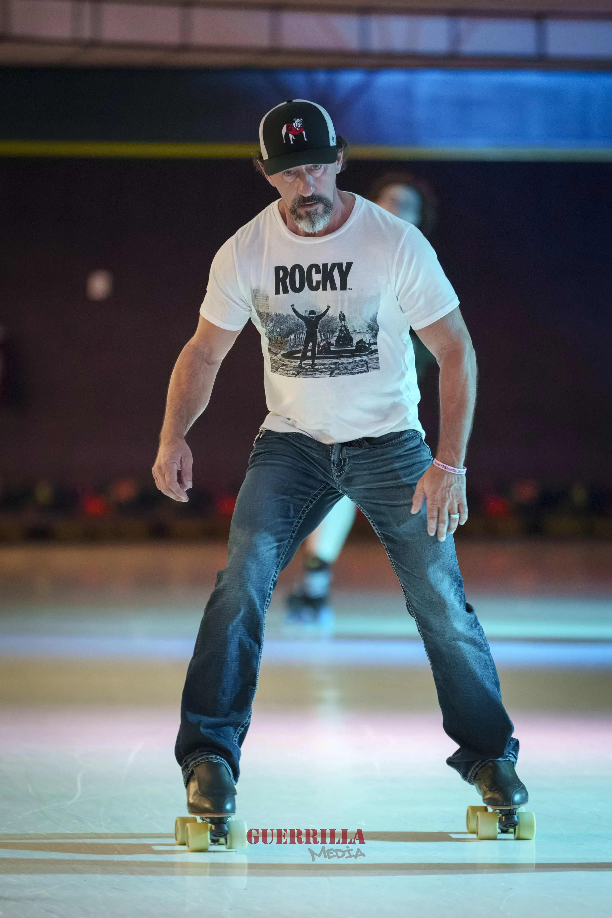 A man wearing a black cap, a white T-shirt with the word 'ROCKY' and a graphic of a person with arms raised, jeans, and roller skates, is skating on a smooth surface in an indoor setting with colorful lighting.