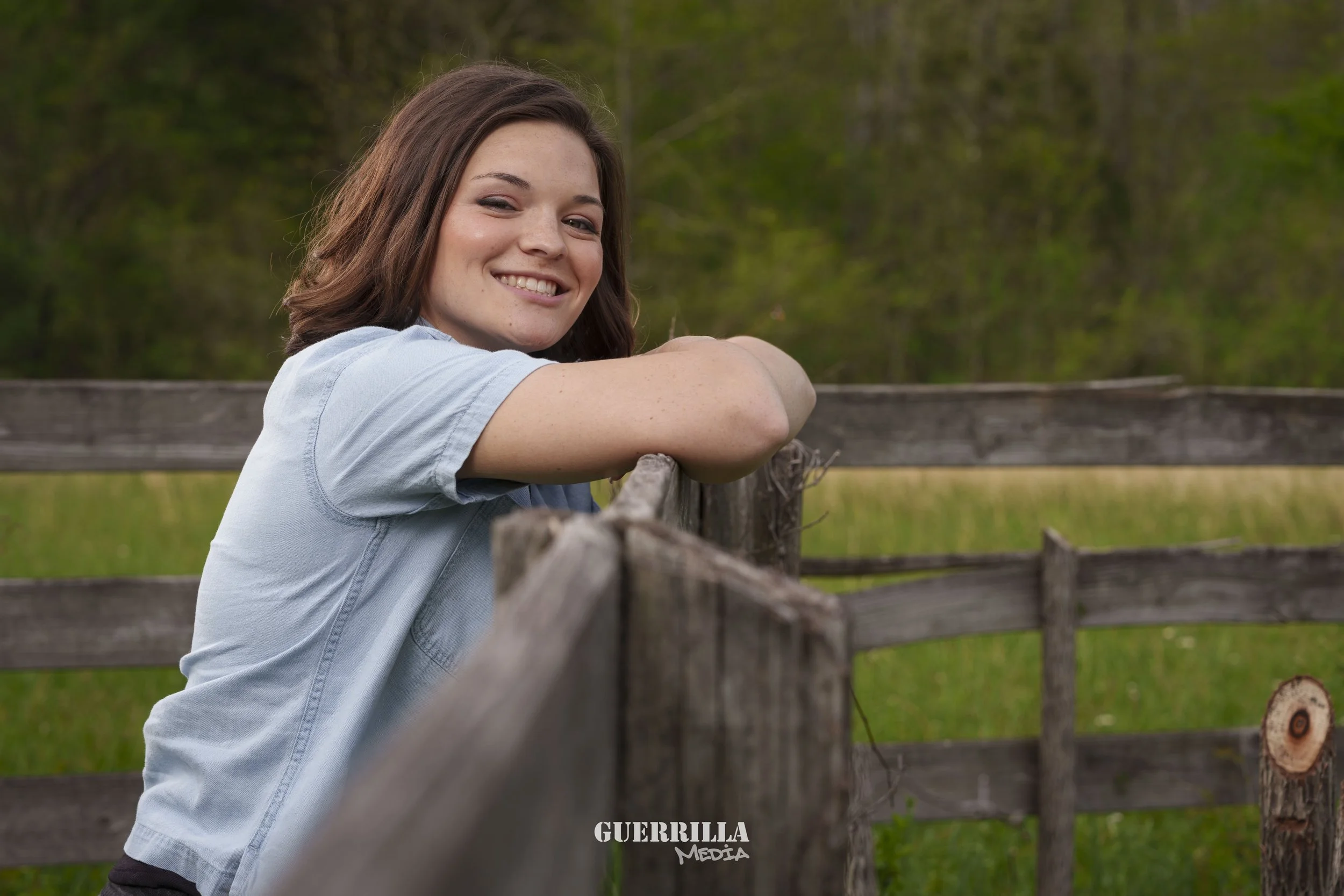 A young woman with shoulder-length brown hair smiling, leaning on a wooden fence in an outdoor setting with green trees and grass in the background.