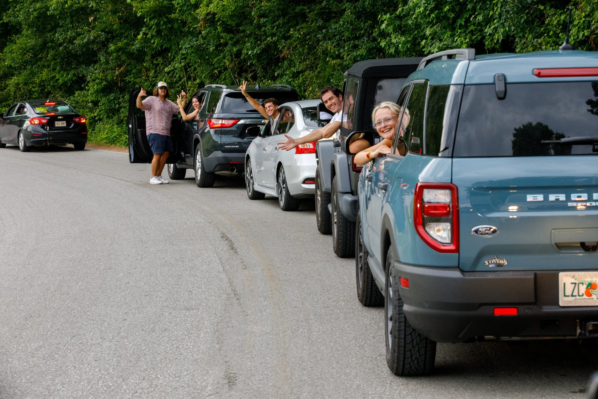 A group of people enjoying a roadside gathering beside several parked cars on a road near a wooded area, with some people sticking their heads out of cars and waving.