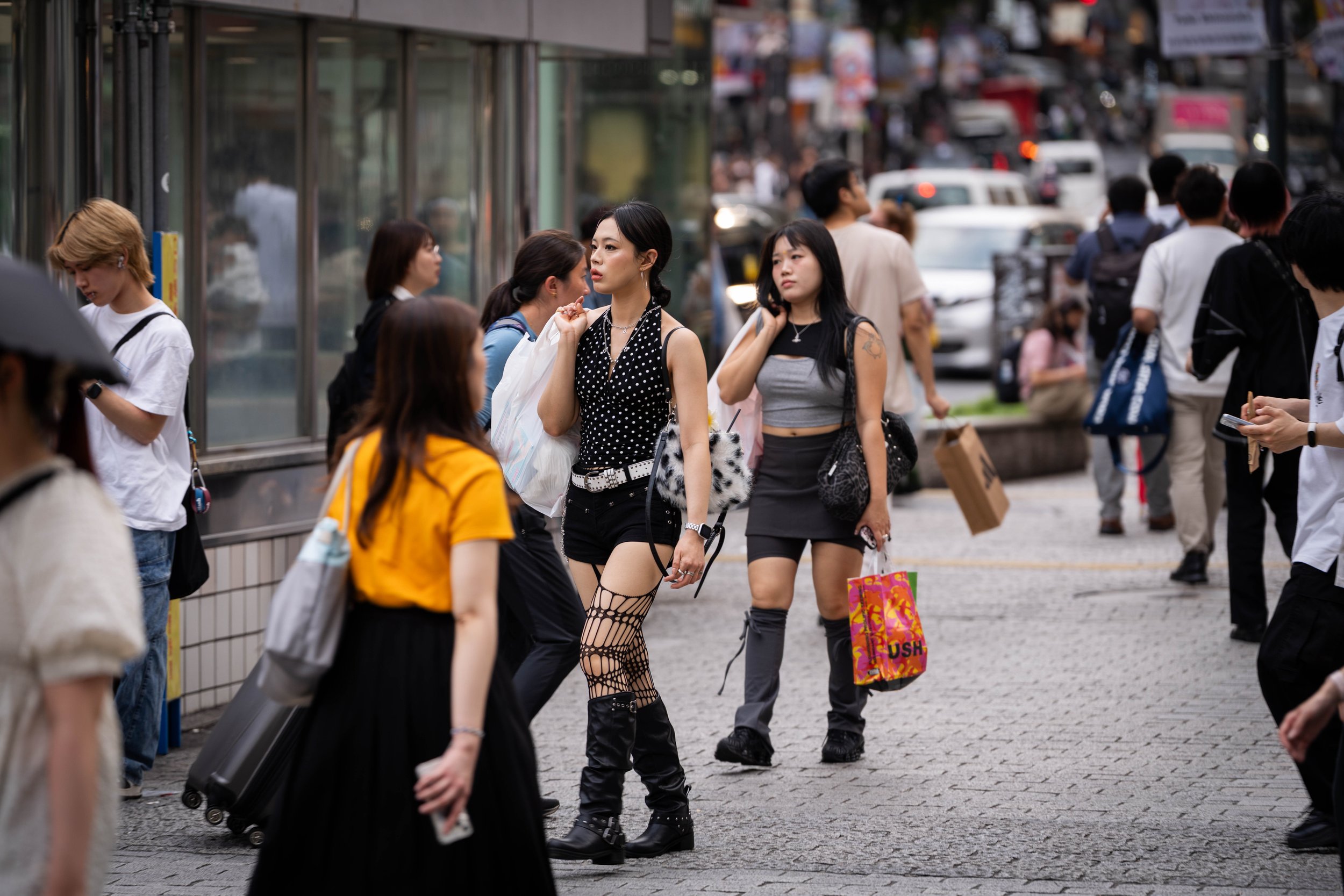 People walking and standing on a crowded city sidewalk, some carrying shopping bags, with vehicles and buildings in the background.
