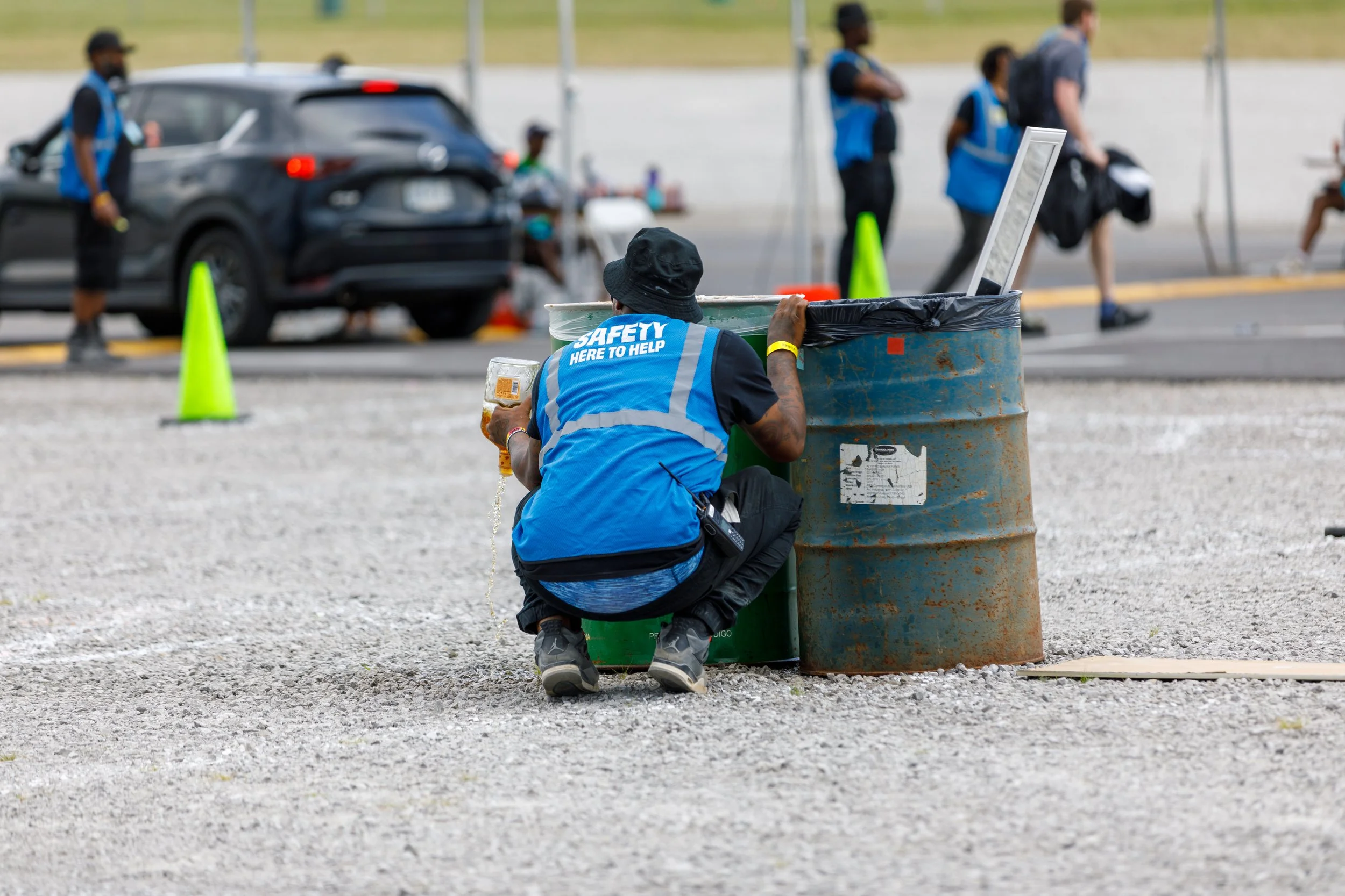 A safety worker kneeling on the ground near a large trash bin, holding a drink in one hand and facing away from the camera, with a computer monitor inside the trash bin. In the background, there are several people and cars at an outdoor event.