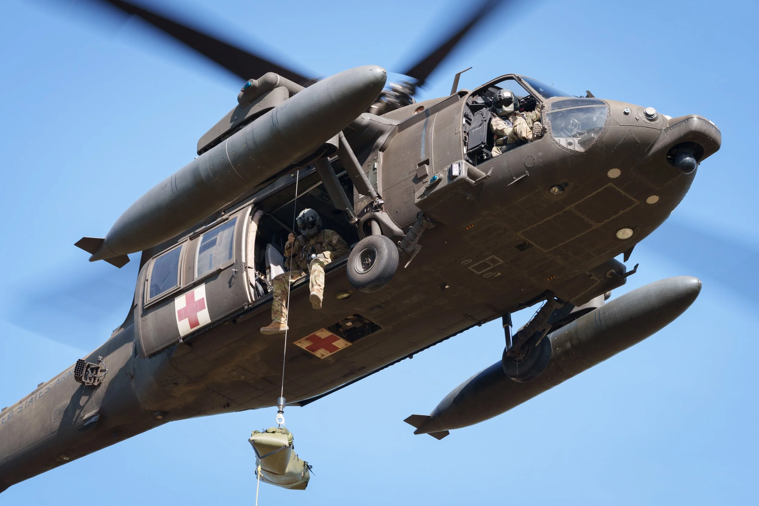 A military medical helicopter in flight with personnel in the open side door and a medic hanging from a rope carrying a medical bag.