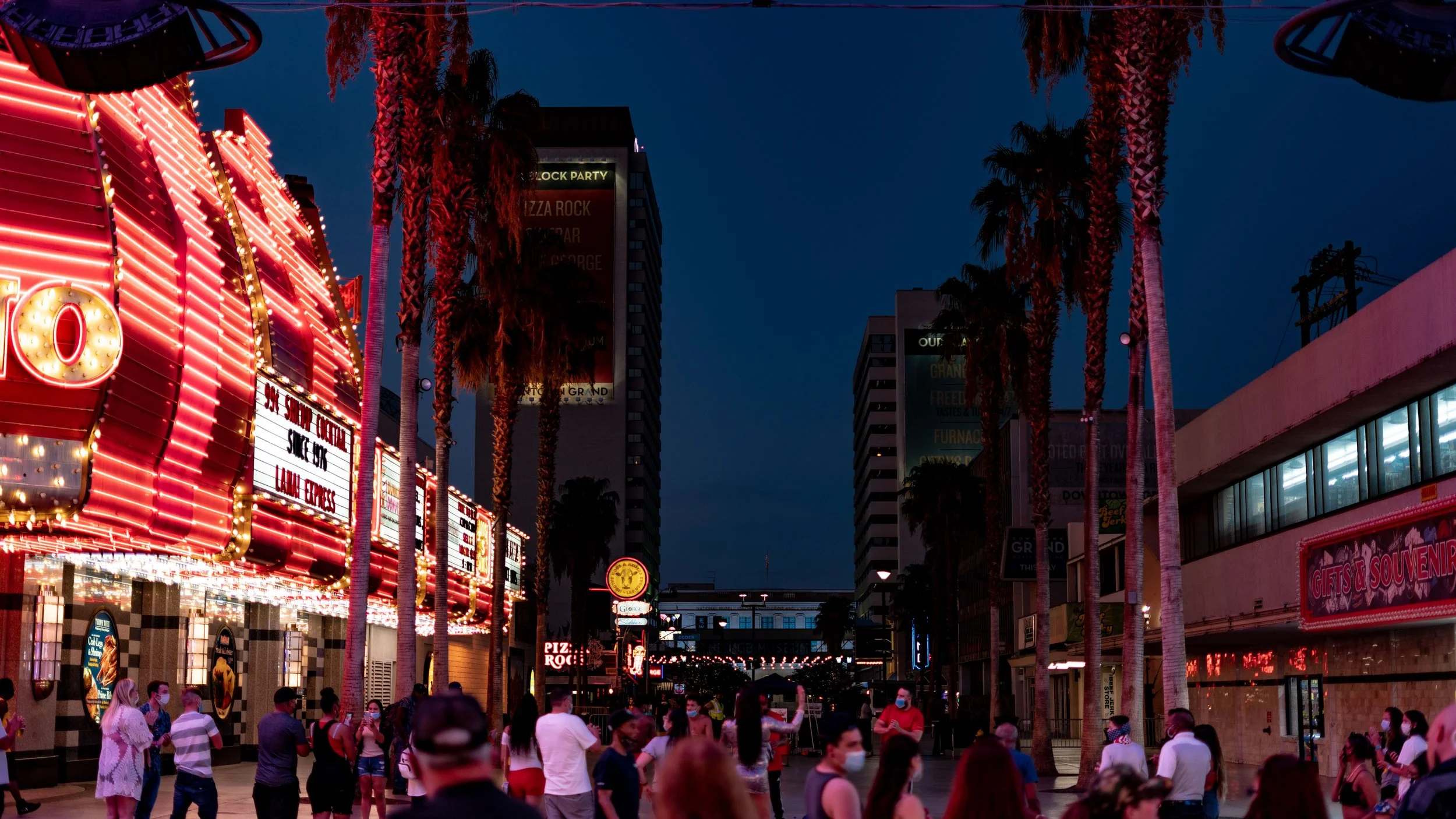 Nighttime street scene with illuminated marquee sign and crowd of people wearing masks, palm trees lining the street, and tall buildings with signs in the background.