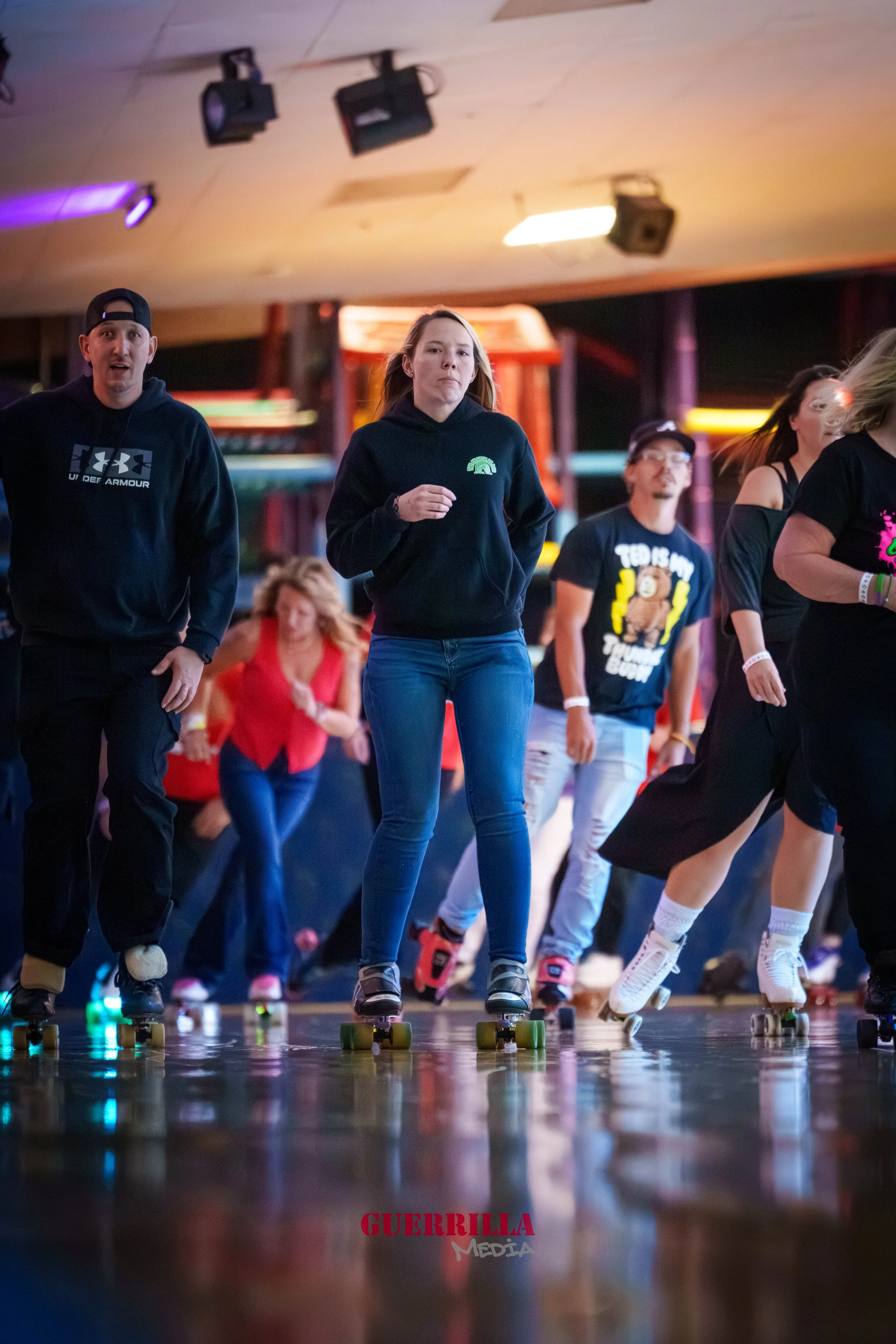People roller skating indoors with a wooden floor, colorful lights, and a ceiling with mounted speakers.
