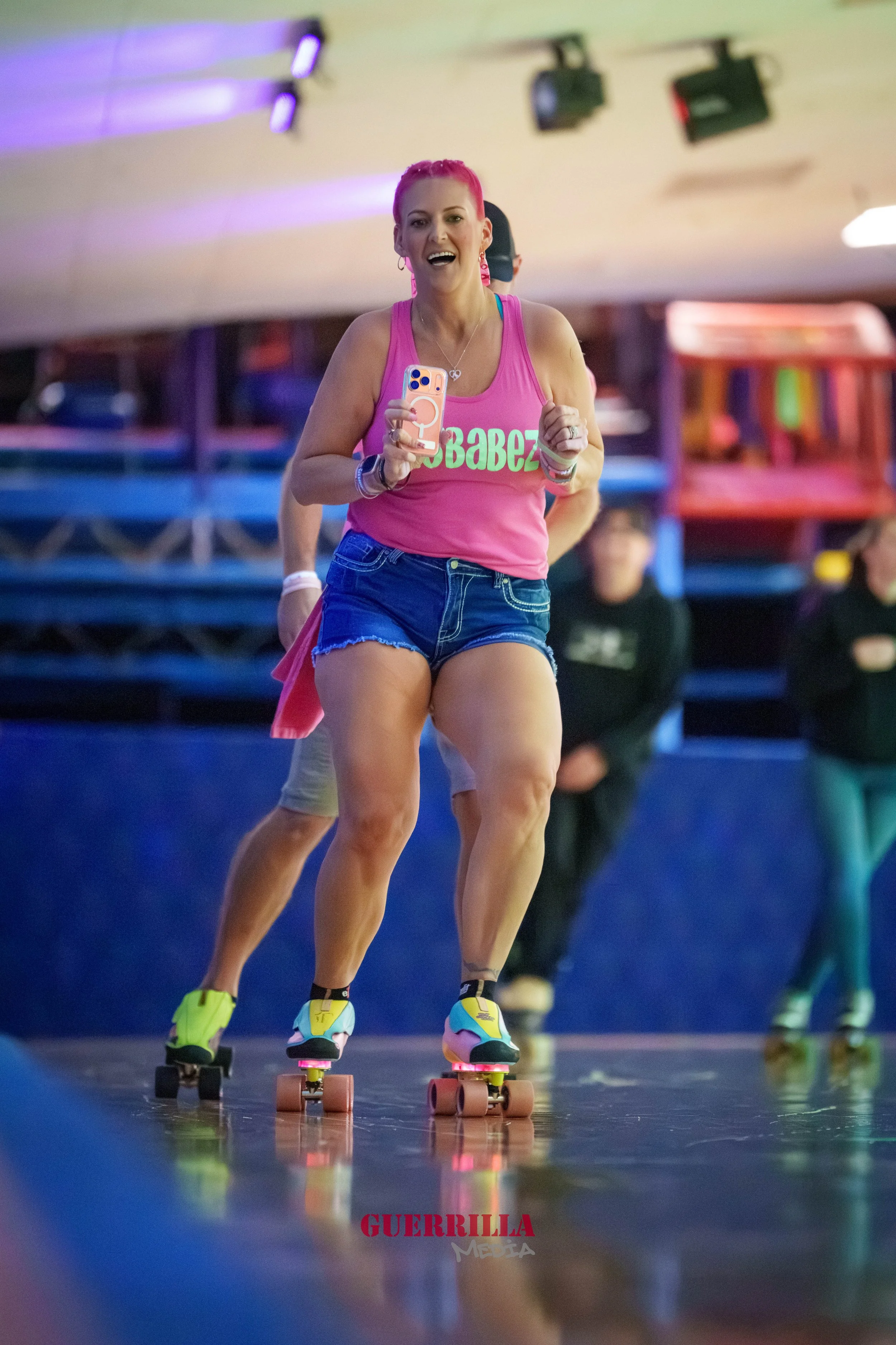 A woman roller skating indoors wearing a pink tank top with green text, denim shorts, and colorful roller skates, smiling and holding a phone.