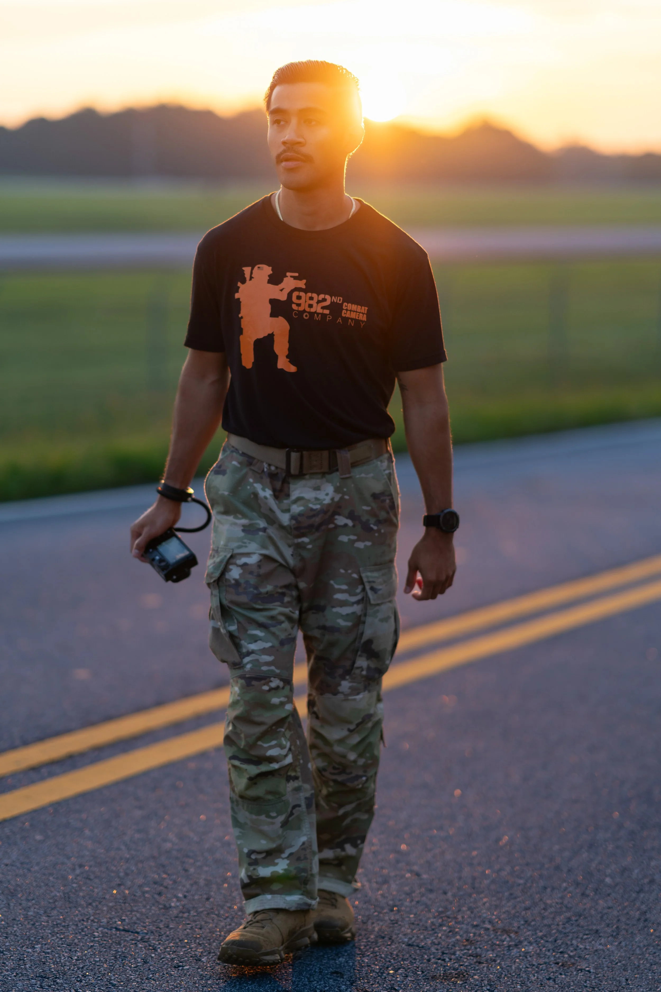 A man walking on a road at sunset, wearing camouflage pants and a black t-shirt with a graphic of a soldier and the text '982nd Combat Company'.
