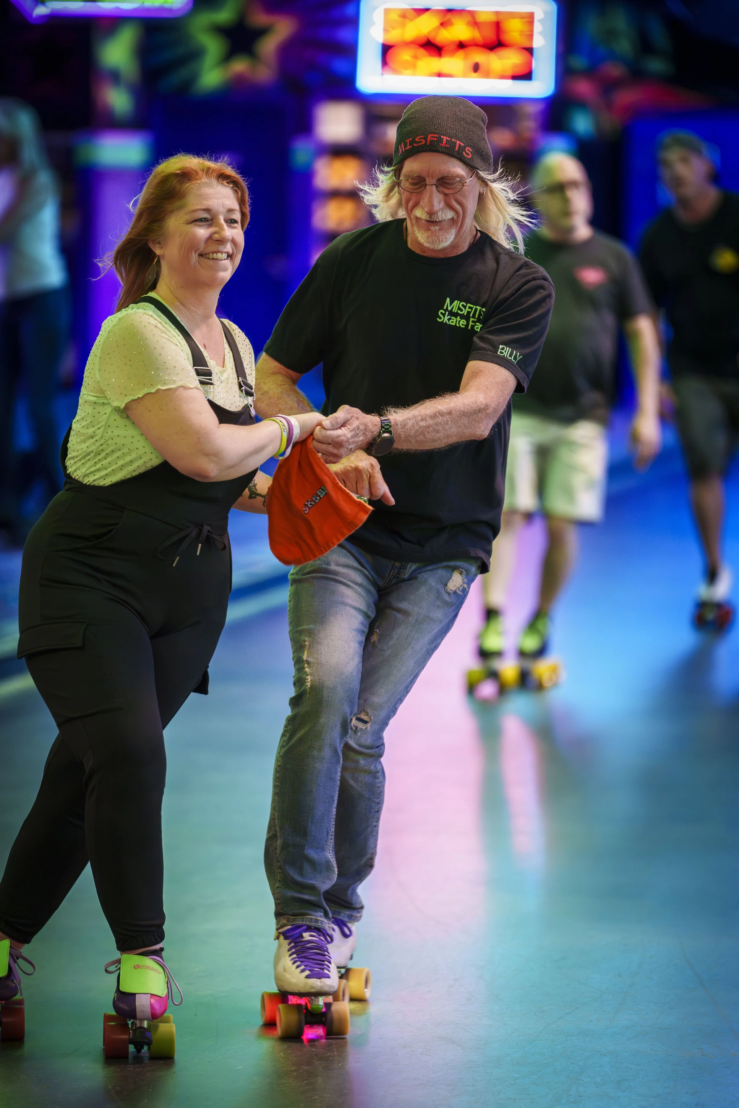 A woman and a man roller skating together in an indoor roller rink with colorful lighting. The woman is smiling and holding the man's hand, and the man is wearing a black beanie and saying 'MISFITS' on his shirt.