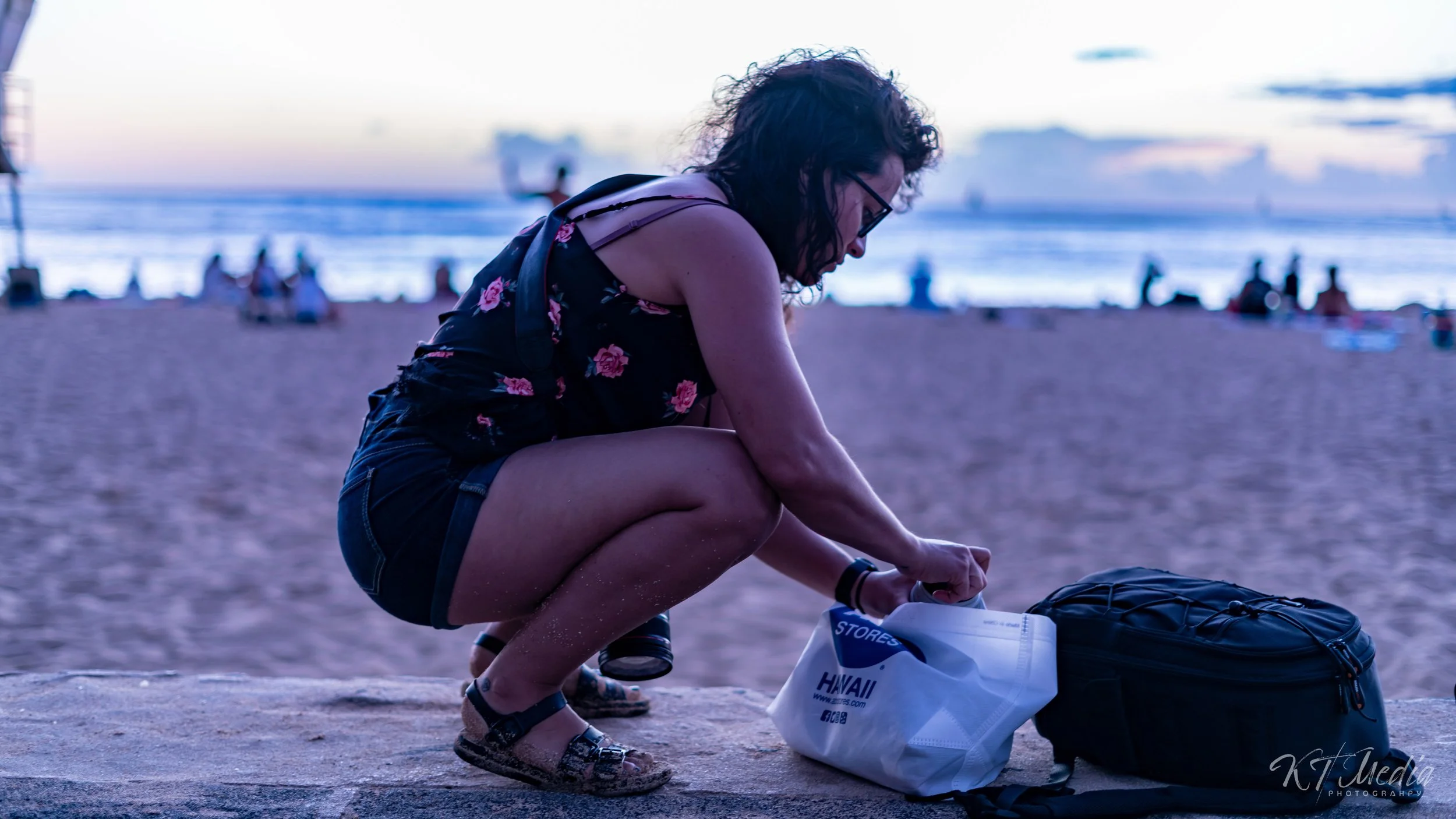 Woman crouching on a beach at sunset, packing a white bag labeled Hawaii with a black backpack next to her.