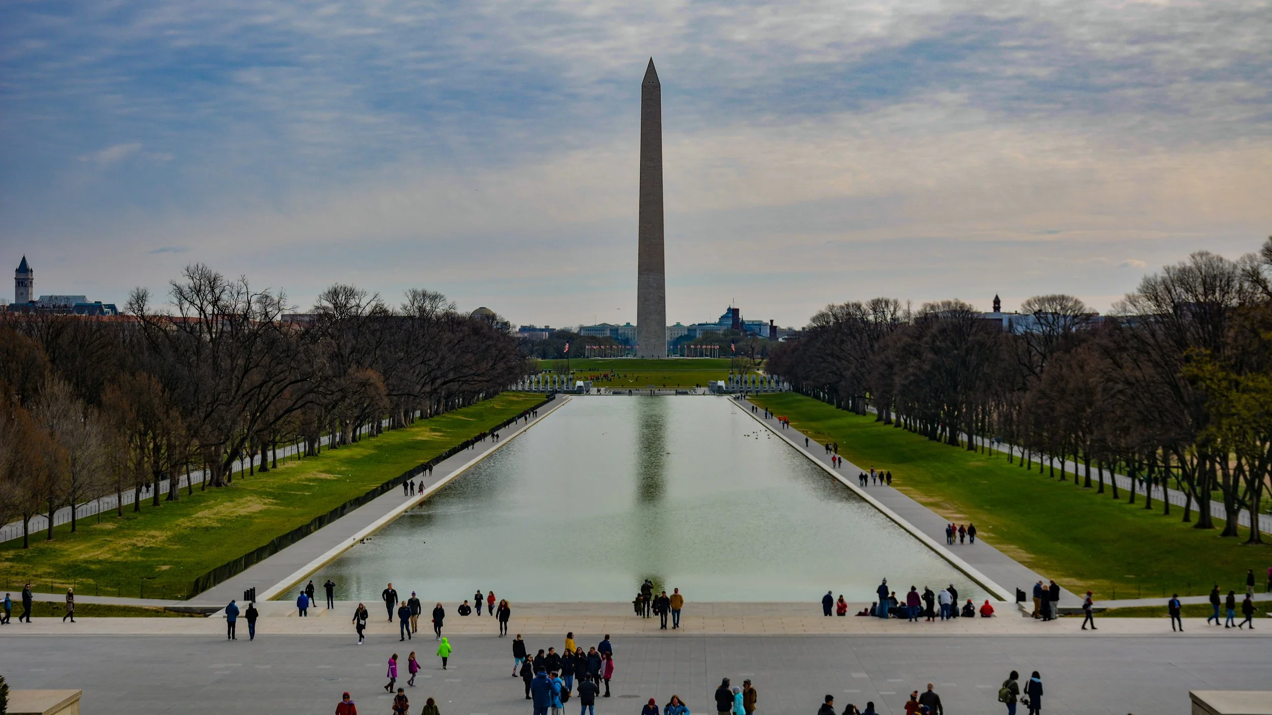 The Washington Monument is in the distance, with the Reflecting Pool in front. People are walking and gathering along the pool and grounds, with leafless trees lining the sides.