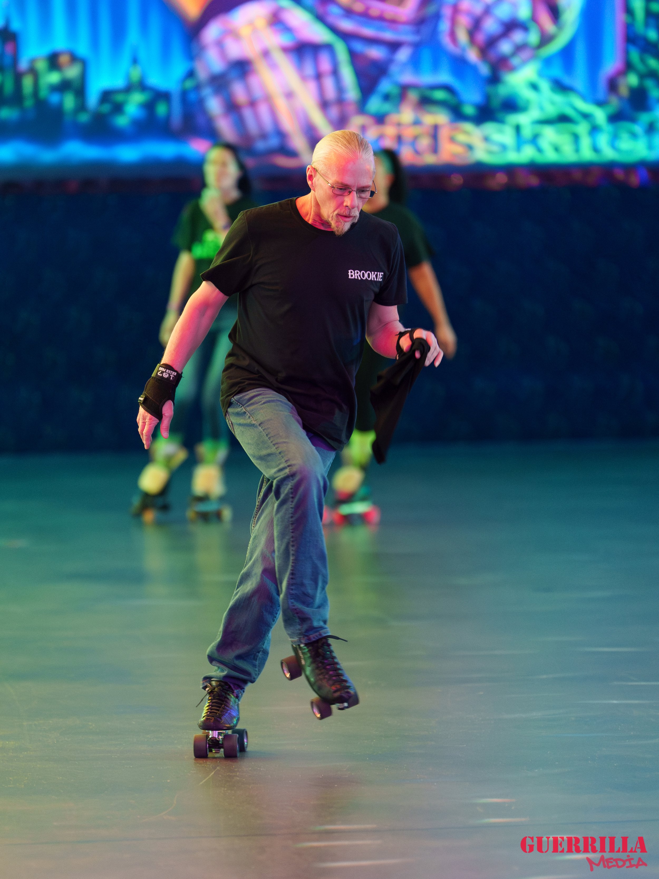 An older man roller skating indoors against a colorful background with others skating behind him.