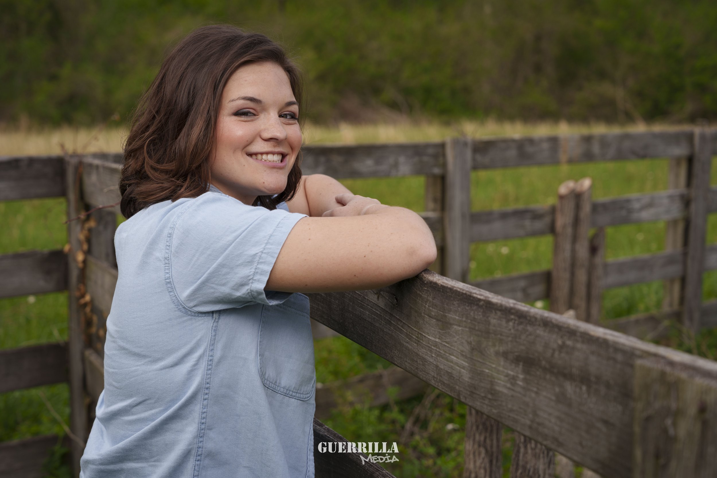 Young woman with shoulder-length brown hair smiling, leaning on a wooden fence in an outdoor field with green grass and trees in the background.