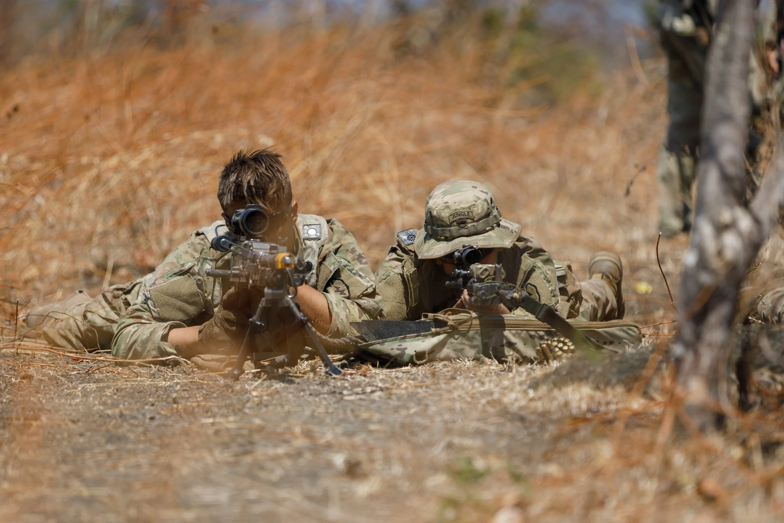 Two soldiers lying on the ground in camouflage uniforms, aiming their rifles while in a wooded area.