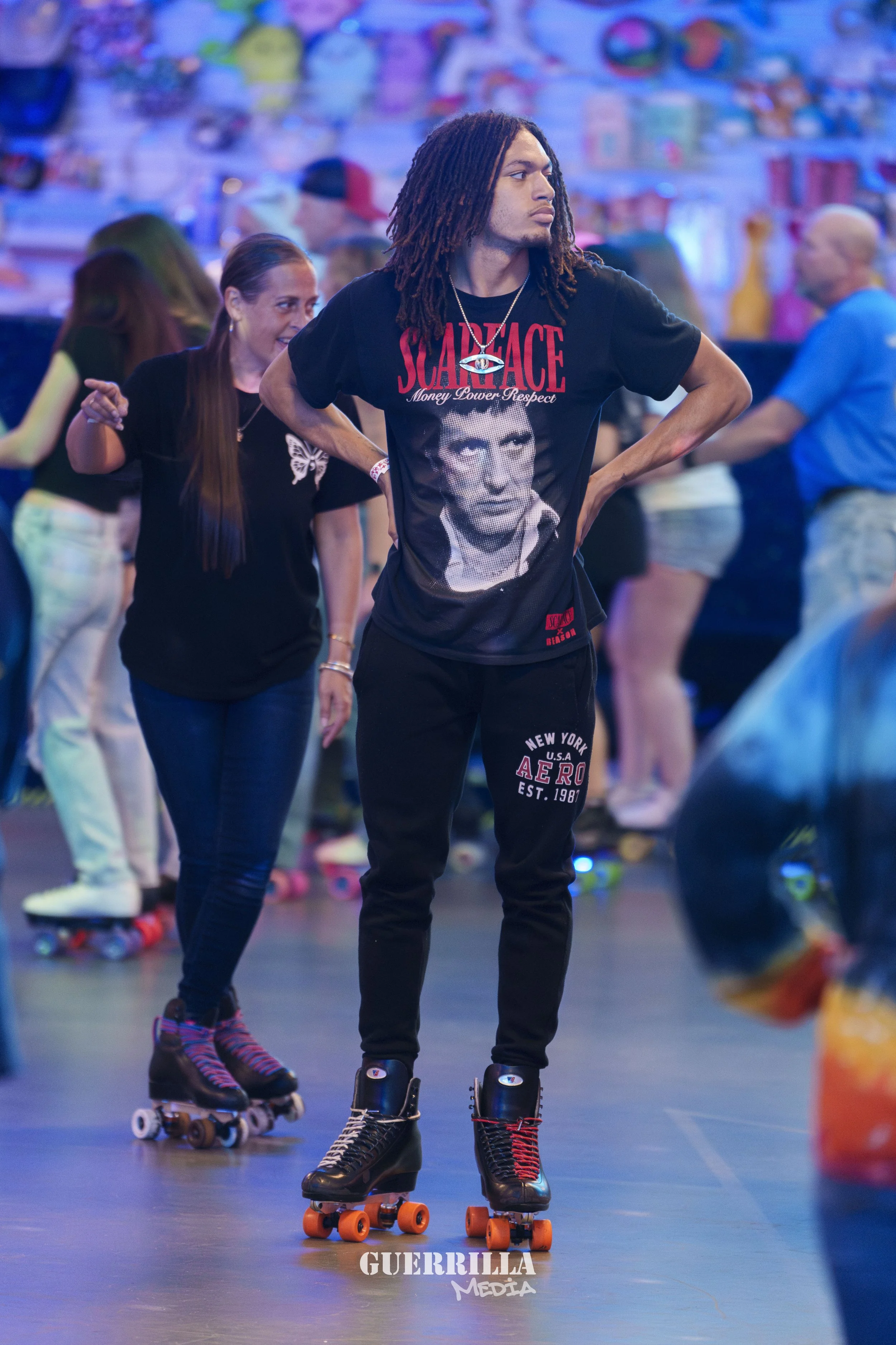 A young man with dreadlocks standing on roller skates in an indoor skating rink, with several other skaters in the background.